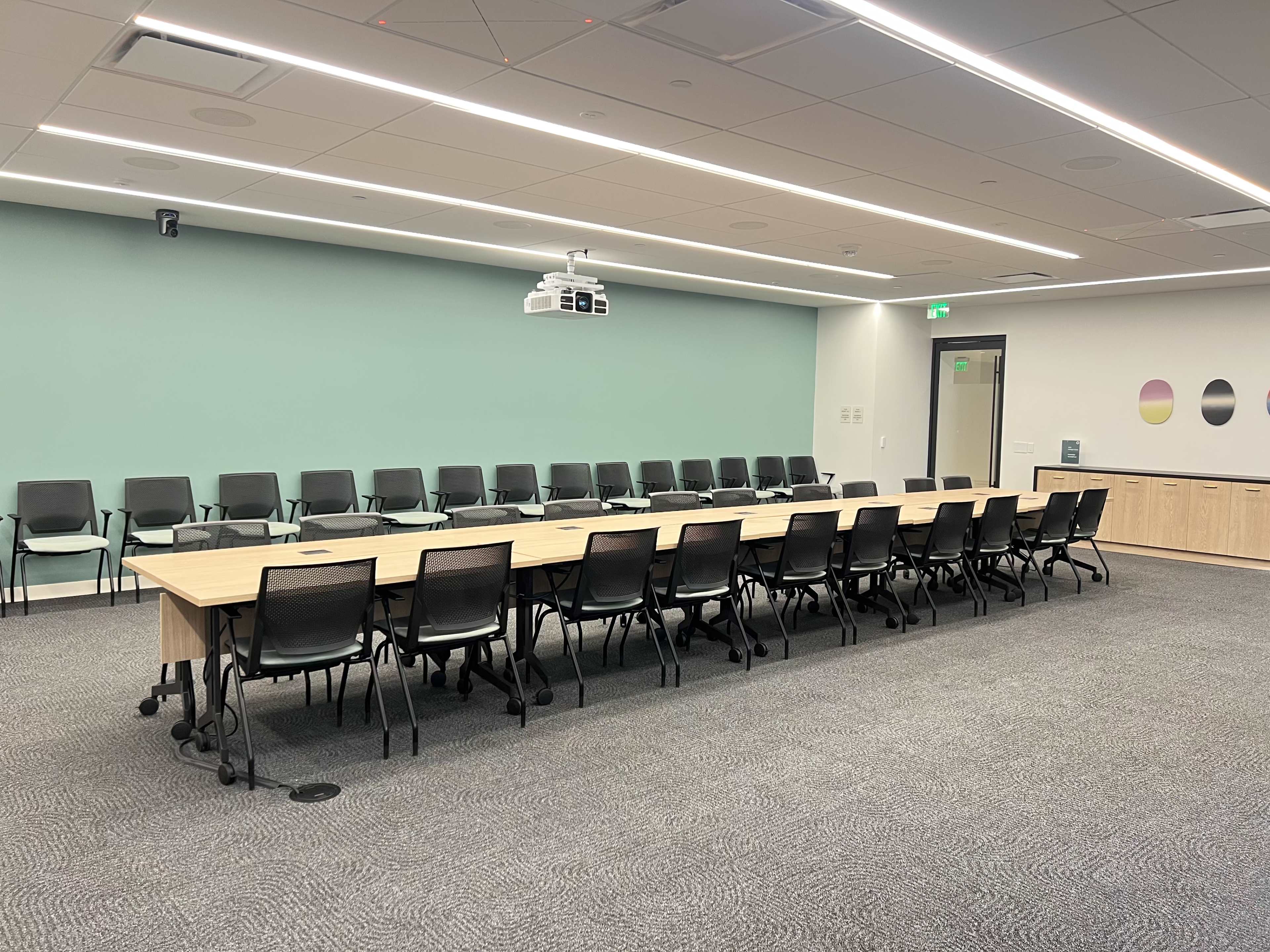 A conference room features a long wooden table surrounded by black chairs, with a projector mounted on the ceiling and a light green wall.