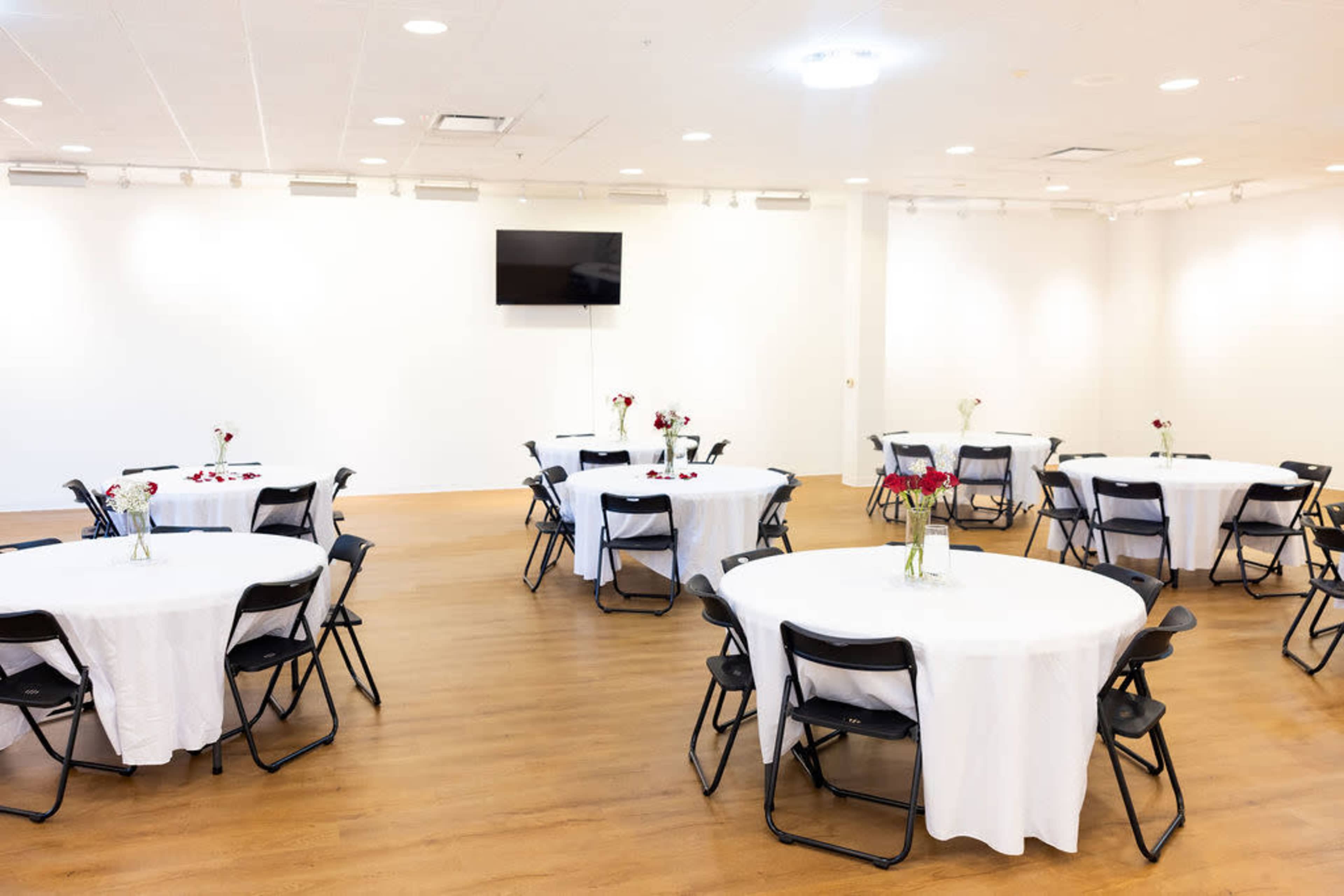 The image shows a spacious indoor event venue with several round tables covered in white tablecloths, each decorated with small vases of flowers, and a large wall-mounted screen in the background.
