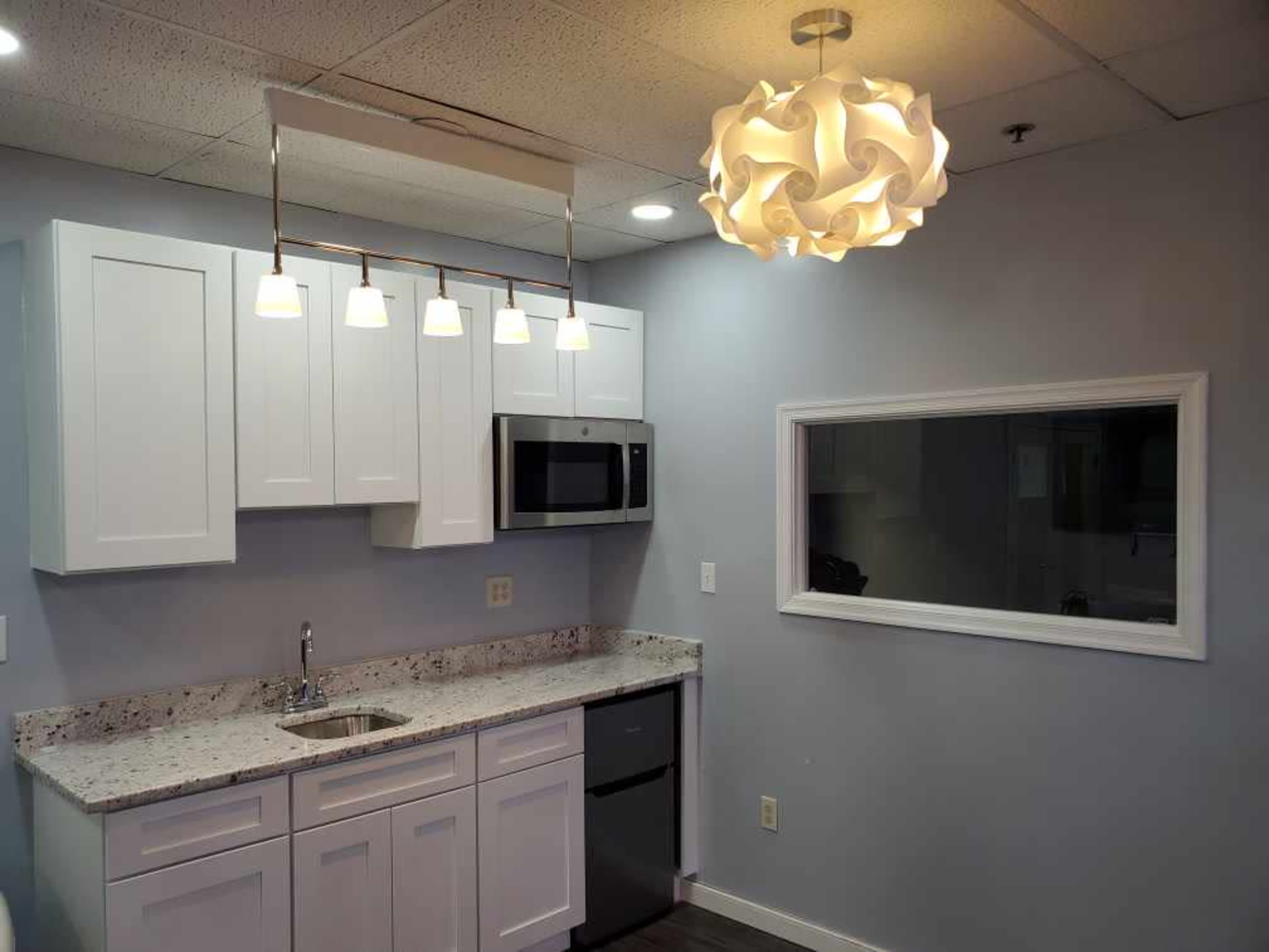 A modern kitchen with white cabinets, a microwave, a stainless steel refrigerator, a sink, and a unique light fixture overhead.
