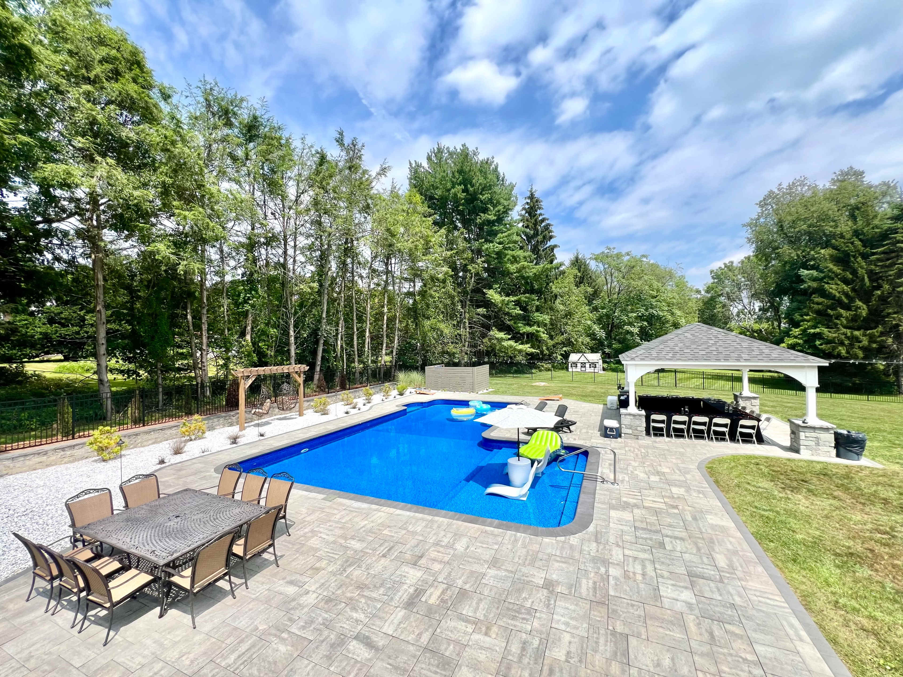 The image shows a backyard featuring a rectangular swimming pool surrounded by lounge chairs, a dining set, and a gazebo.