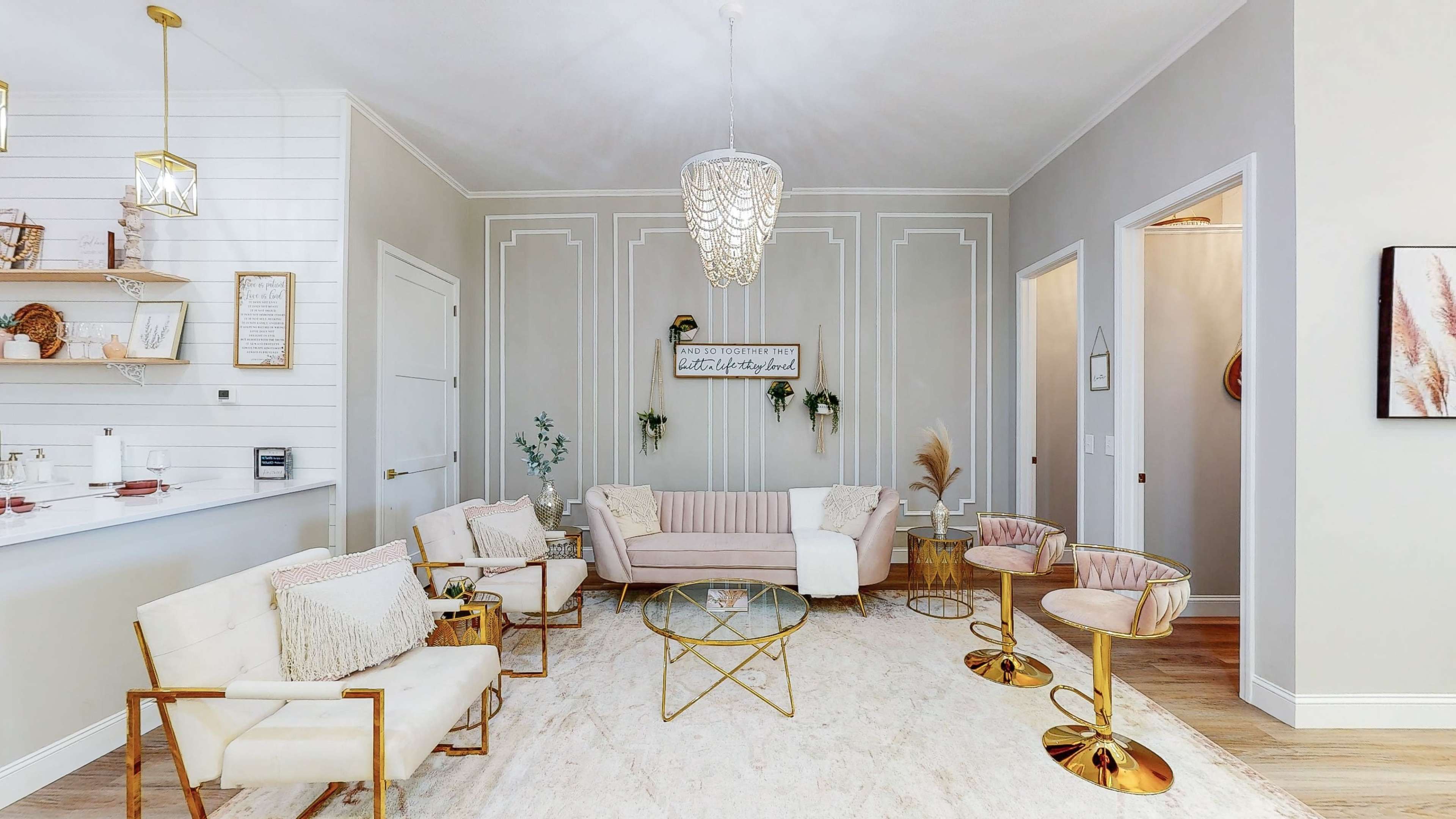 A modern living room features a light-colored sofa and armchairs arranged around a glass coffee table on a large area rug, with decorative wall accents and pendant lighting.