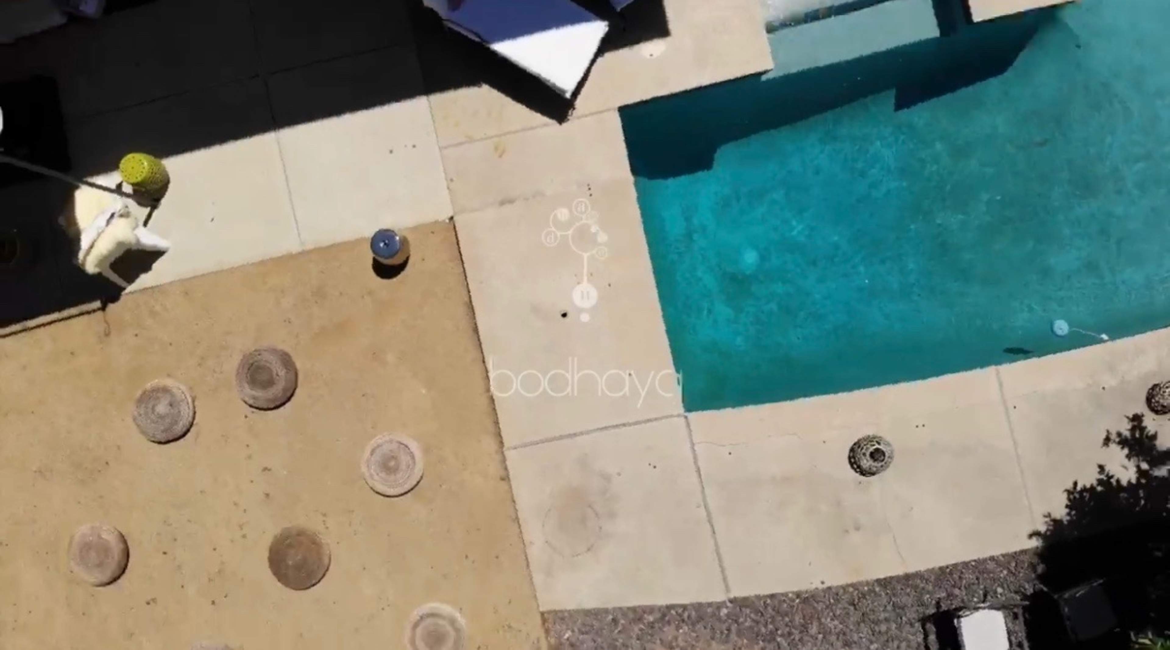 An aerial view of a backyard featuring a pool, patio area with circular stone seating, and surrounding landscaping.