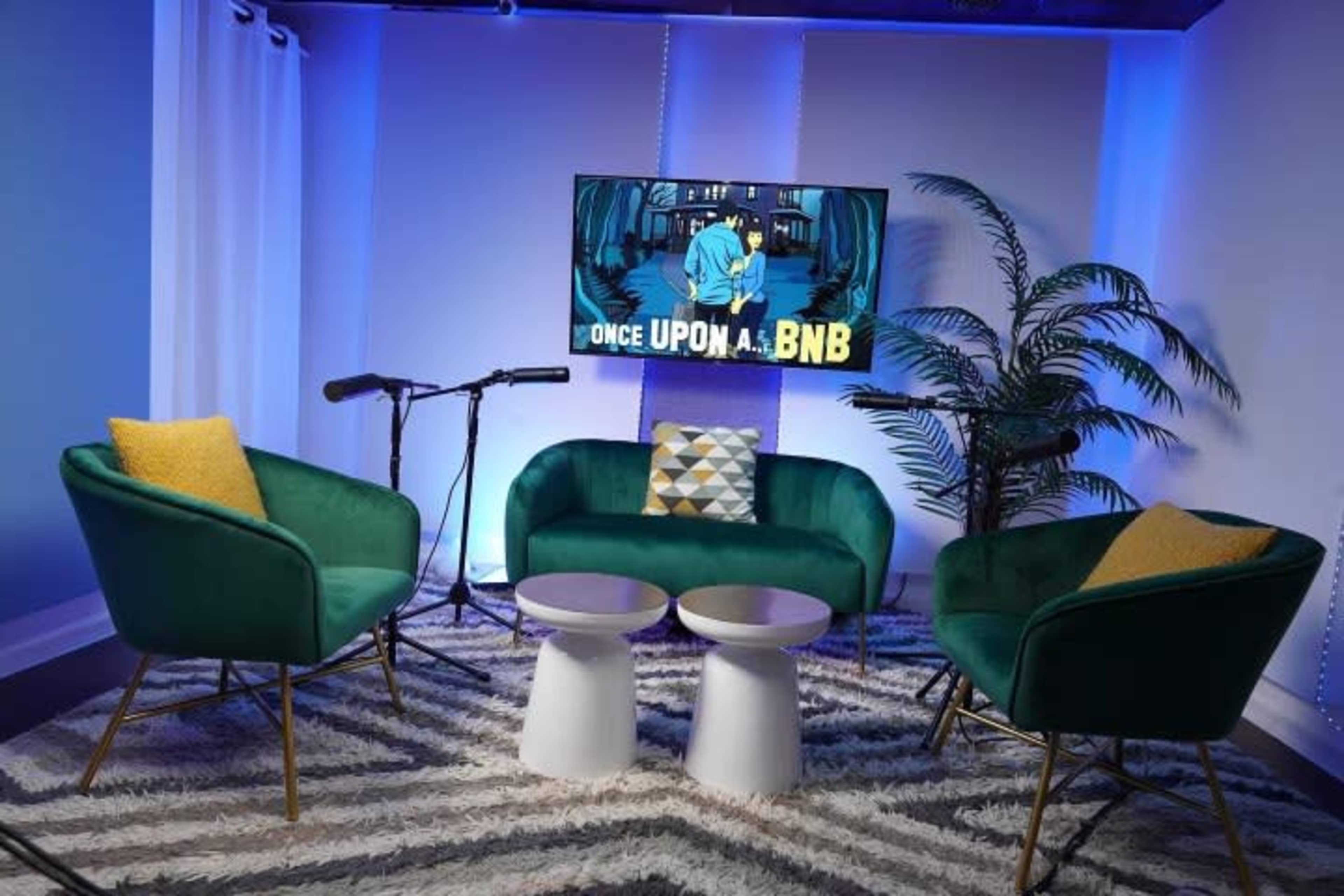 A modern living space features two green chairs and a teal sofa arranged around two white tables, with a large screen displaying "ONCE UPON A . . . BNB" set against a blue-lit backdrop.