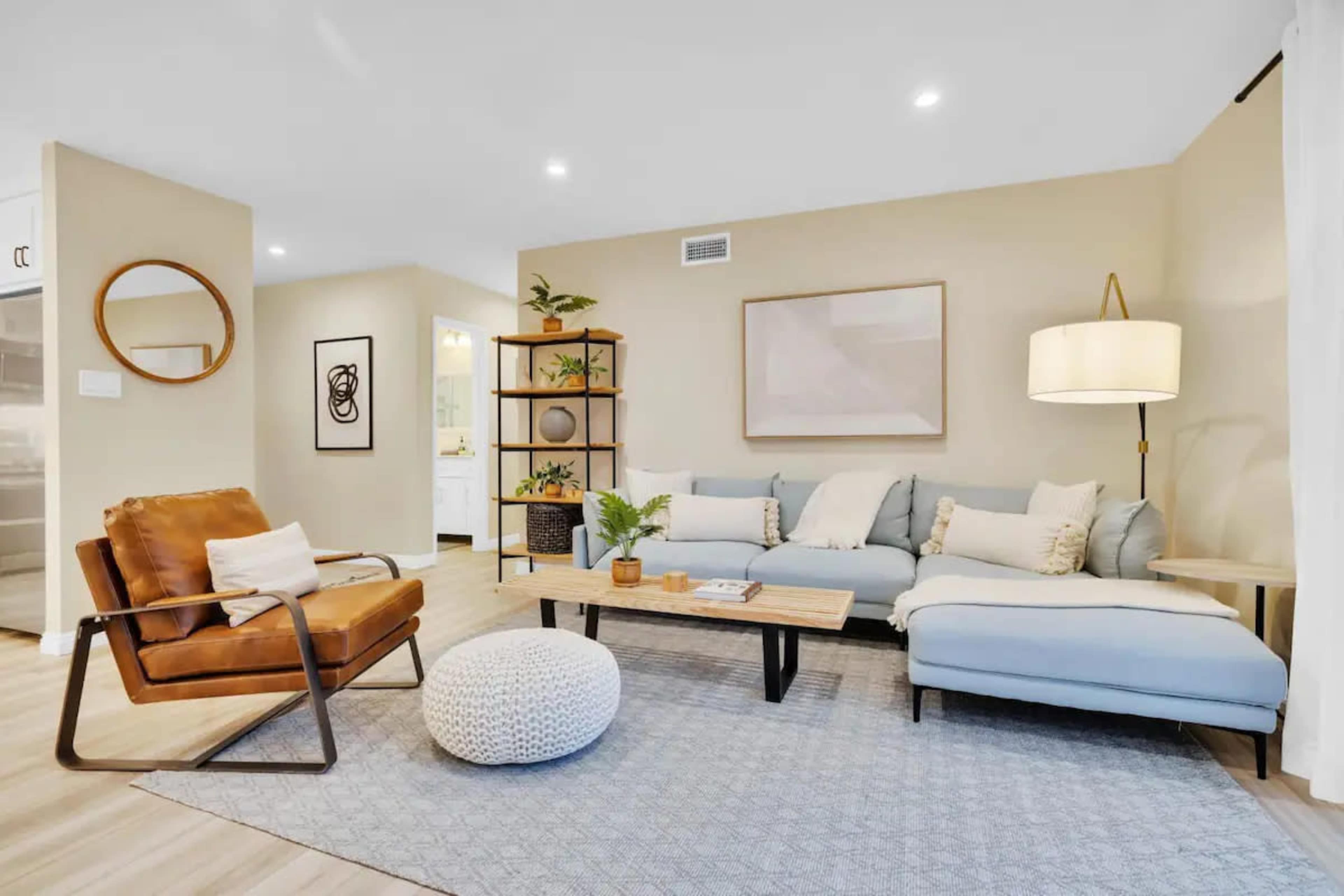 A modern living room with a blue sectional sofa, a light brown leather armchair, a wooden coffee table, and a decorative pouf on a textured area rug.