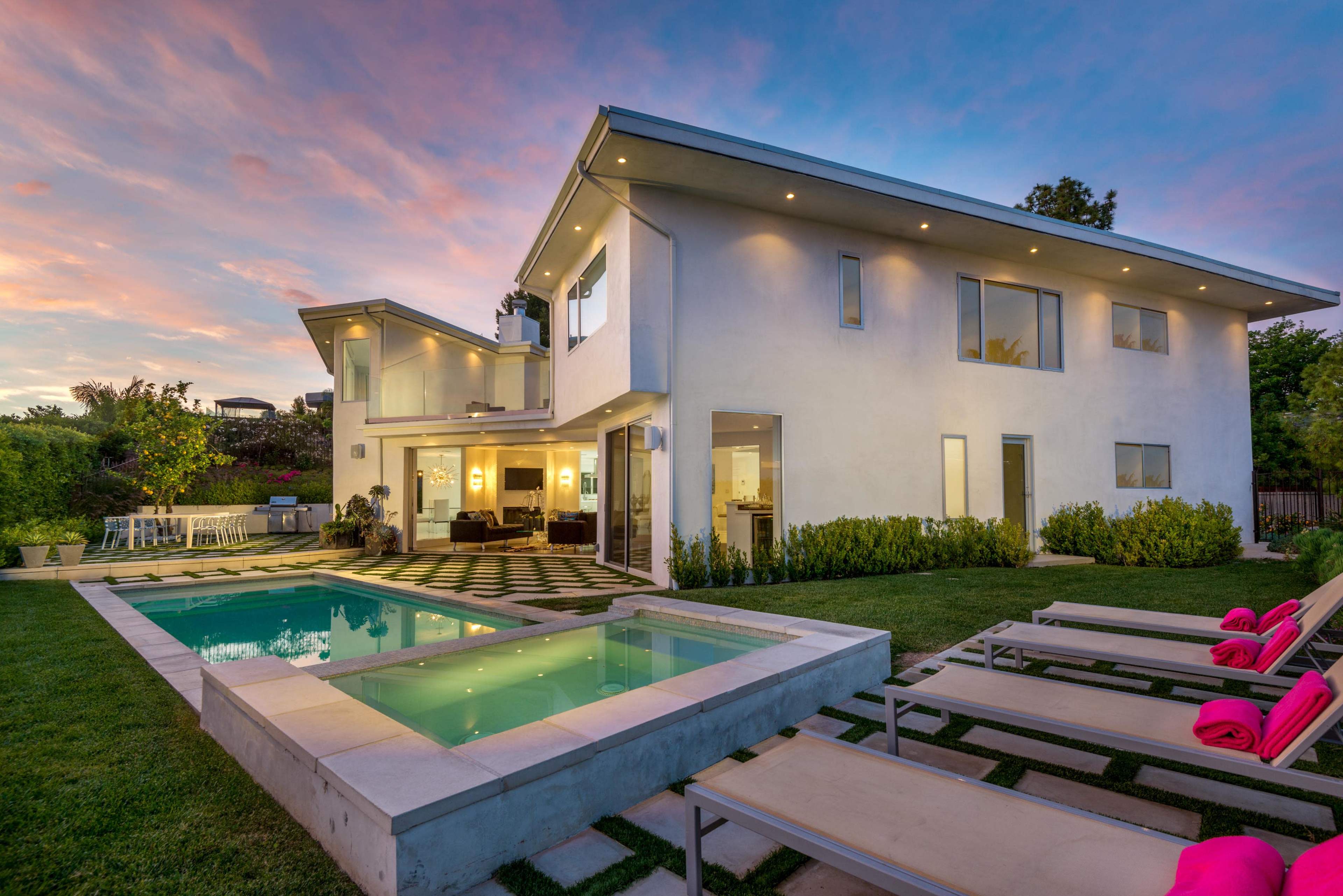 A modern white house with large windows overlooks a pool area surrounded by lounge chairs, set against a colorful sunset sky.