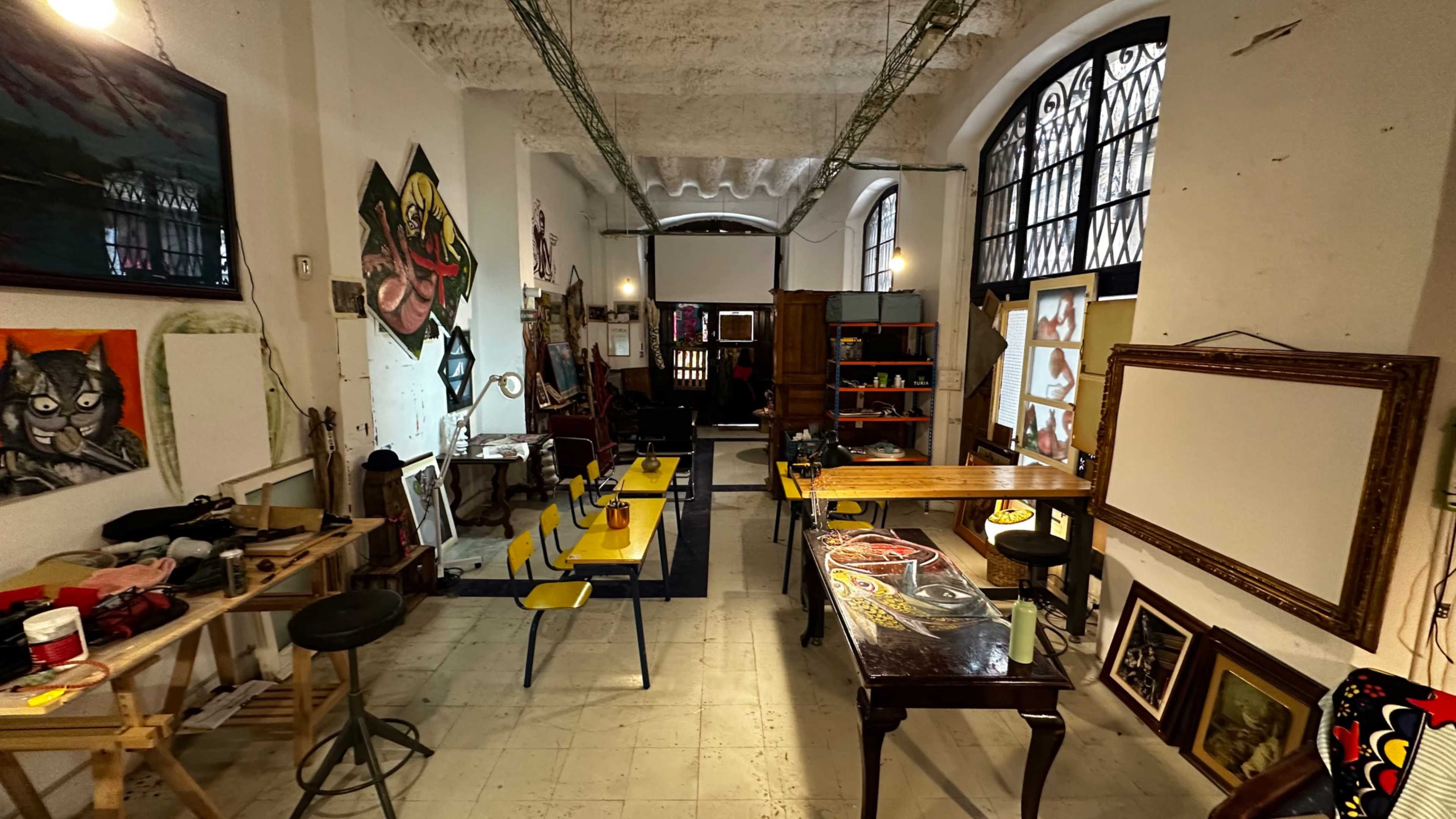 The image shows an artist studio featuring tables with chairs, various artworks on the walls, and a mix of art supplies scattered throughout the space.