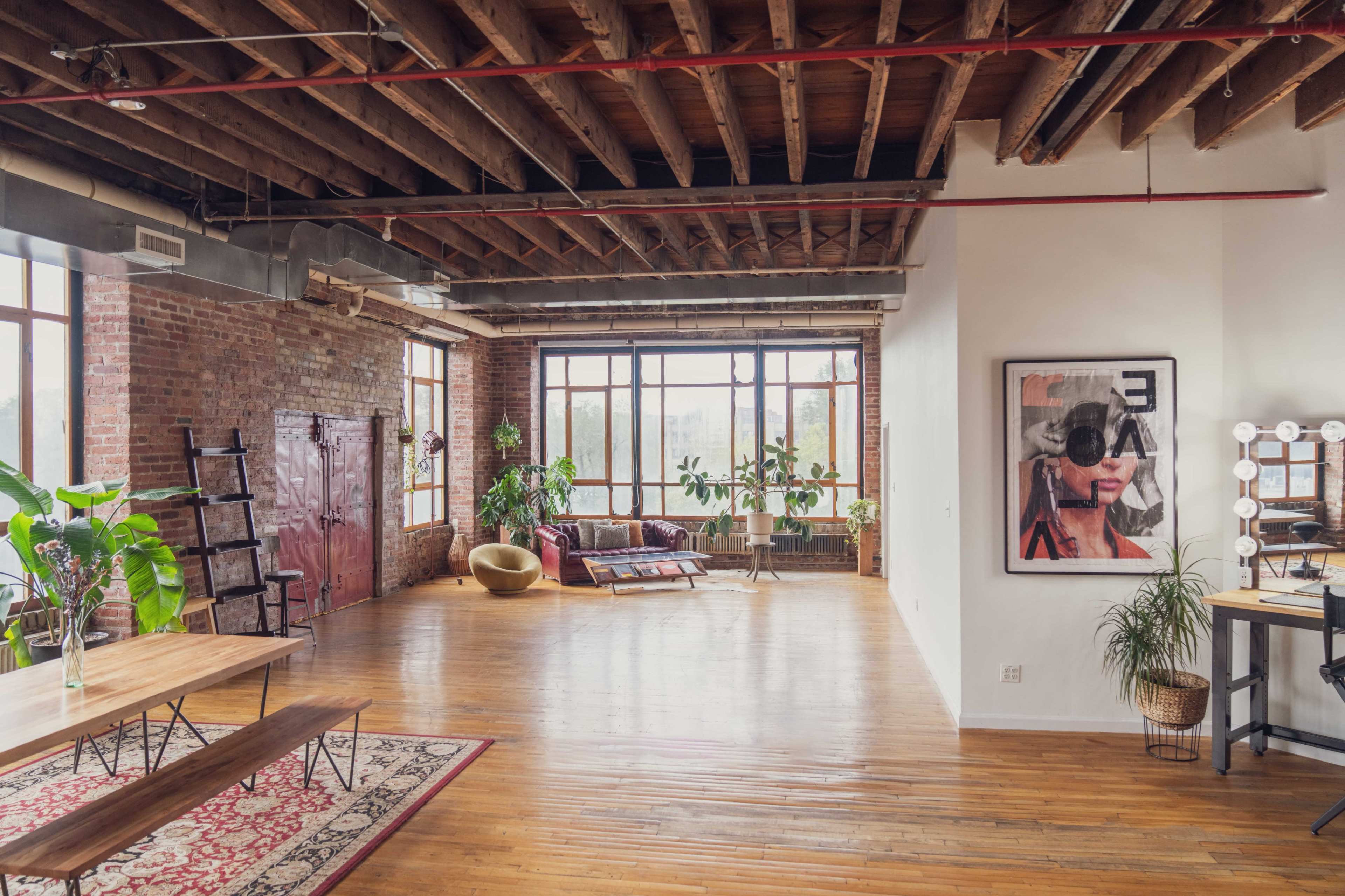 The image shows a spacious industrial-style loft featuring exposed wooden beams, large windows with natural light, and a mix of modern and vintage furnishings.