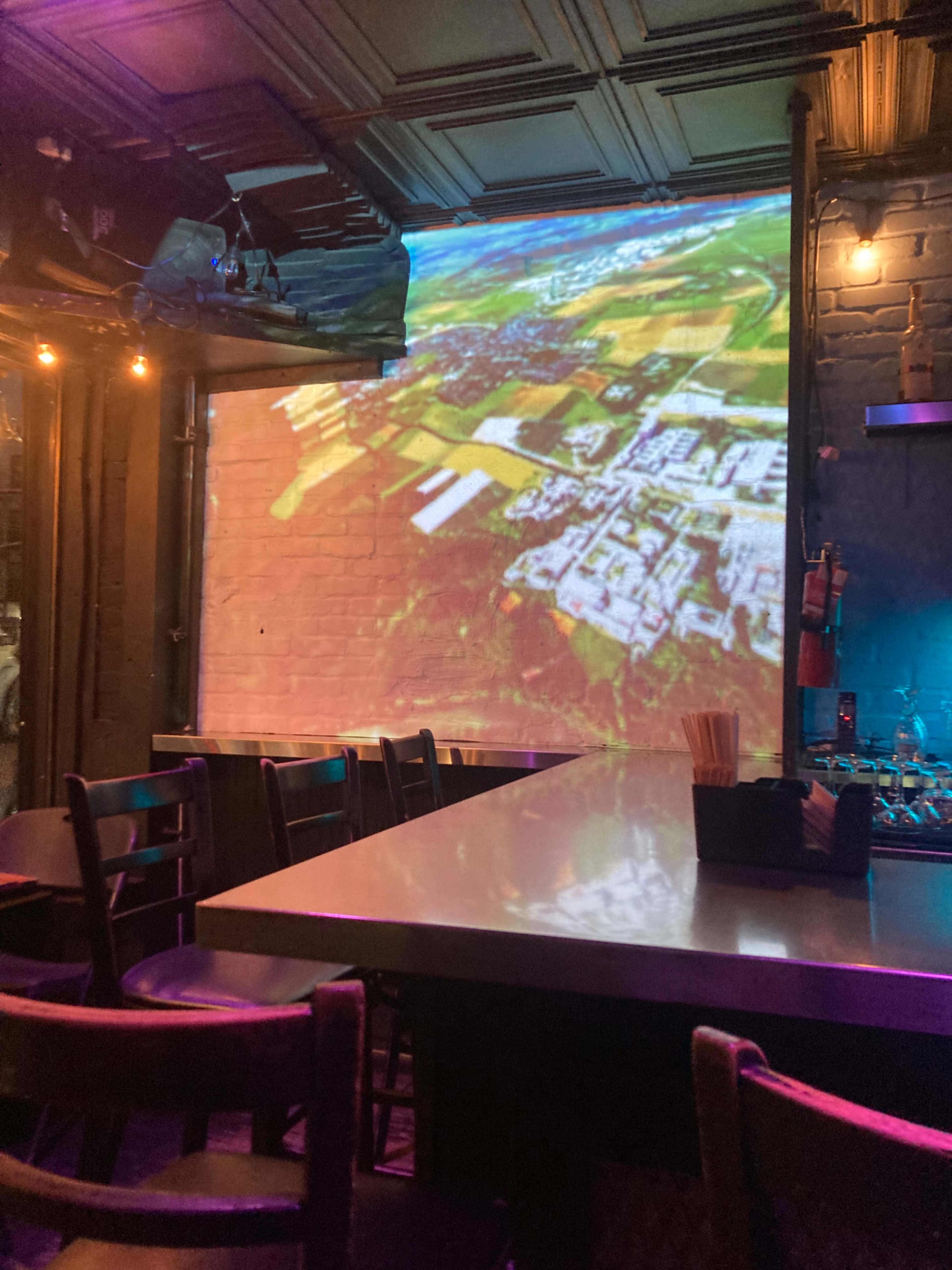 The image shows a bar interior with a projection of an aerial landscape on the wall, while empty chairs and a table are visible in the foreground.
