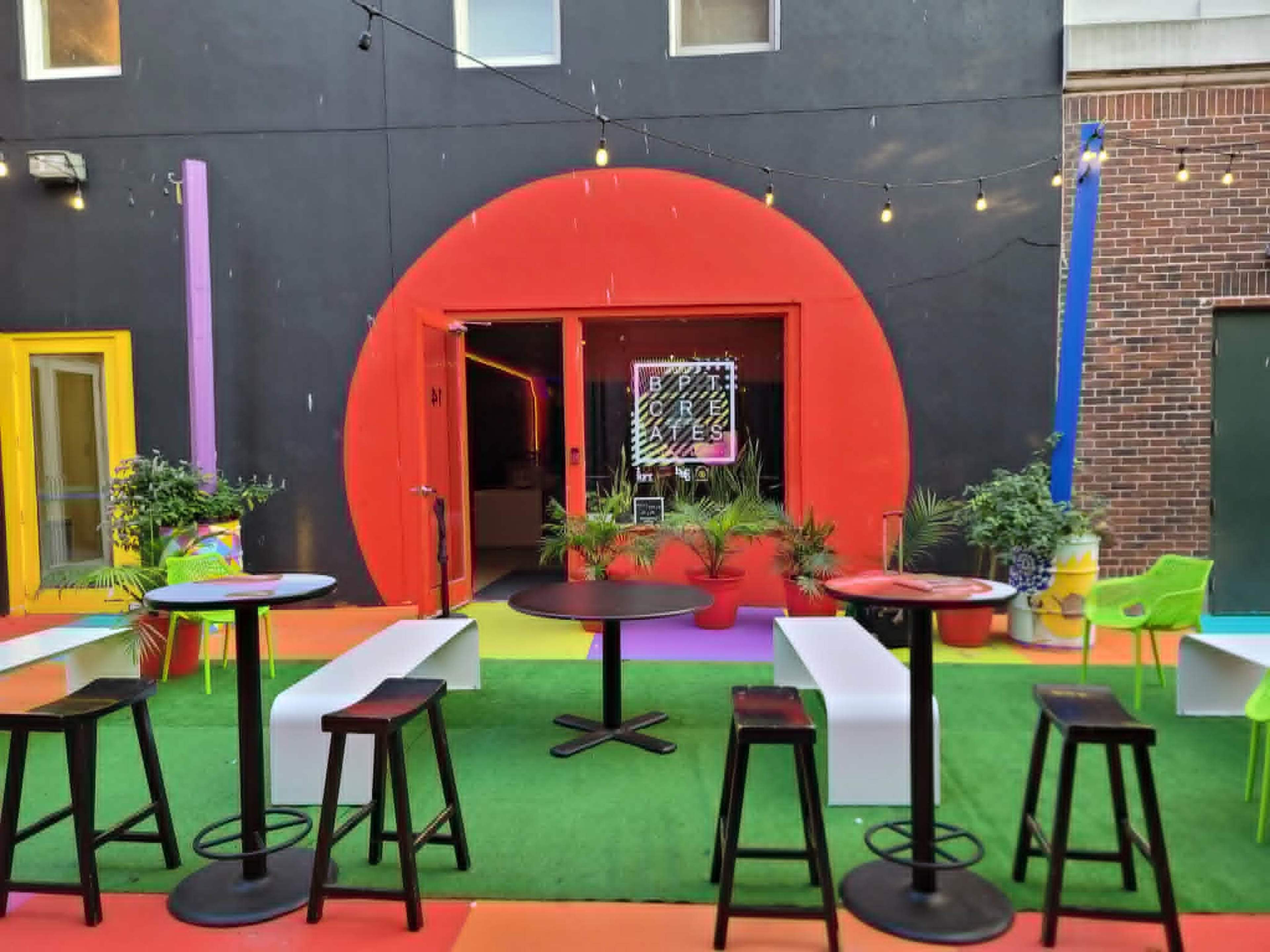 The image shows a vibrant outdoor seating area with colorful furniture and a large circular red entrance to a building in the background.