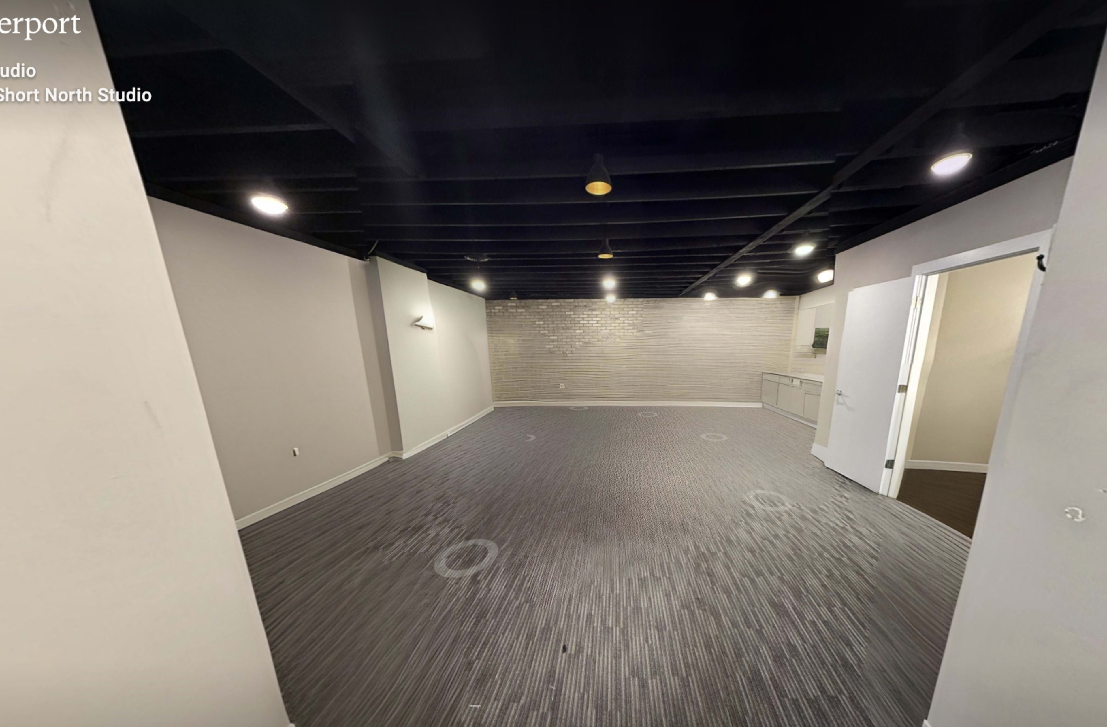 The image shows a spacious, empty studio room with gray walls, a light-colored brick accent wall, and a dark ceiling.