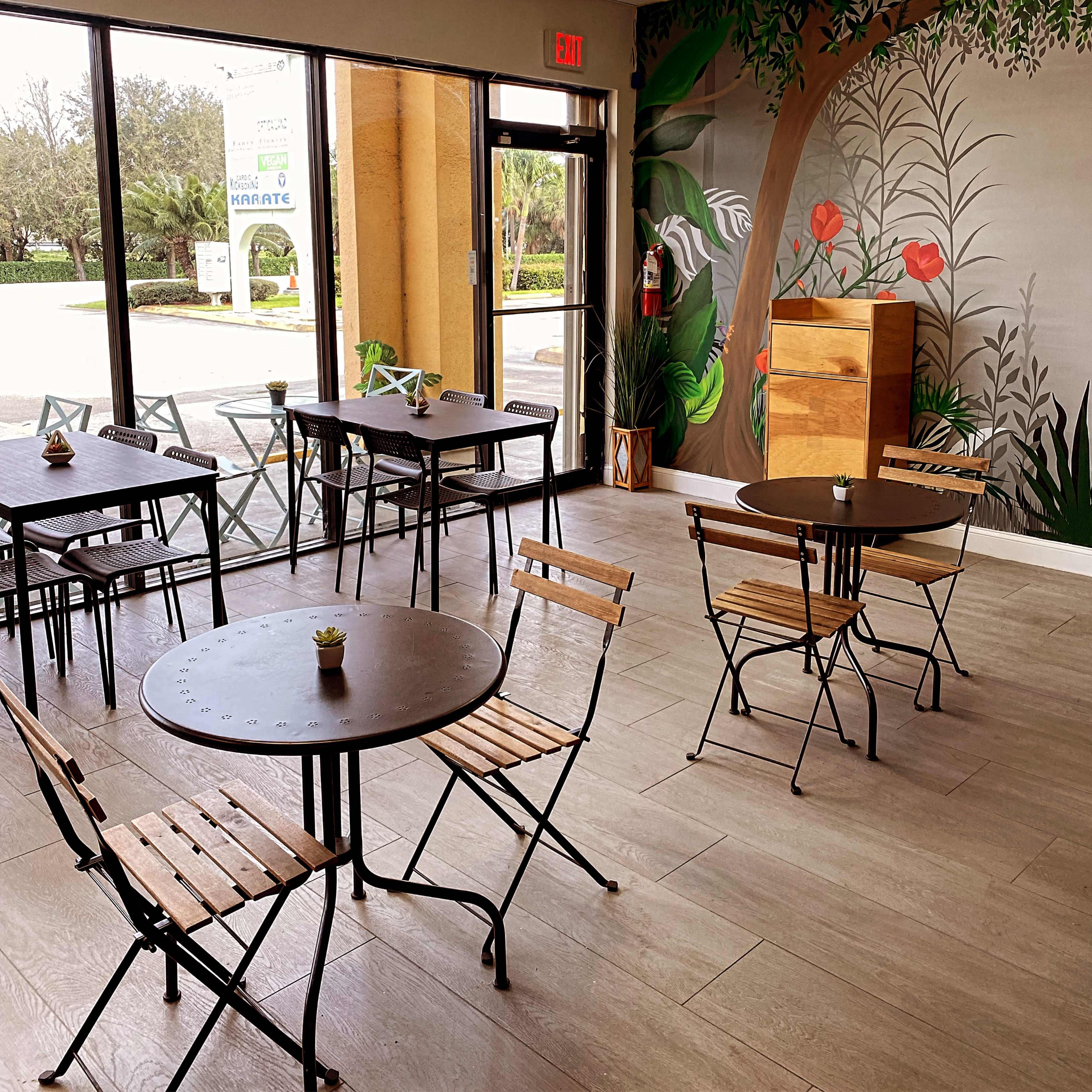 The interior features several black metal tables and wooden chairs arranged in a bright, open space with a mural of greenery on the wall.