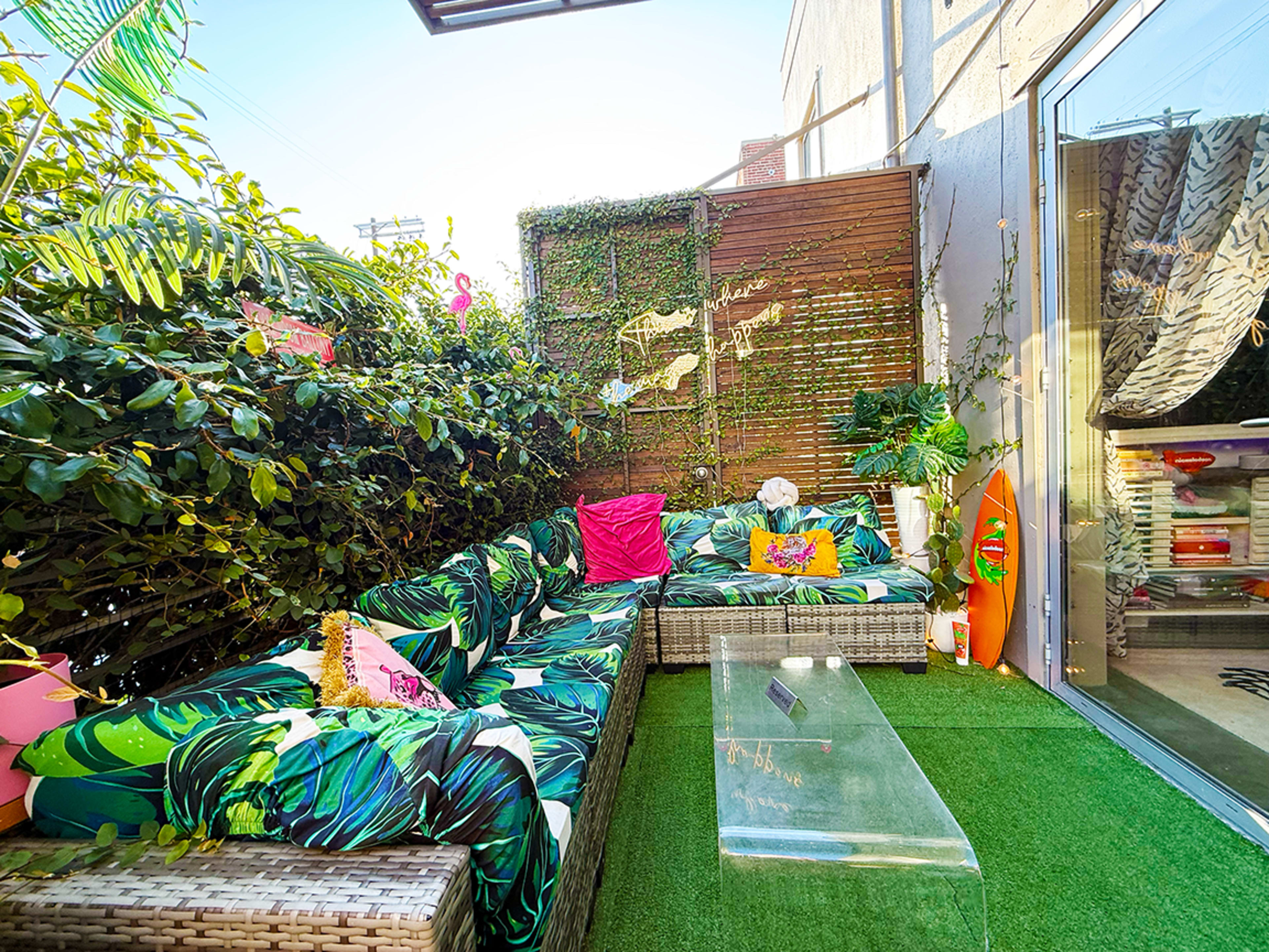 The image shows a cozy outdoor seating area with a green palm leaf-patterned sofa, decorative pillows, and a clear glass coffee table surrounded by greenery.