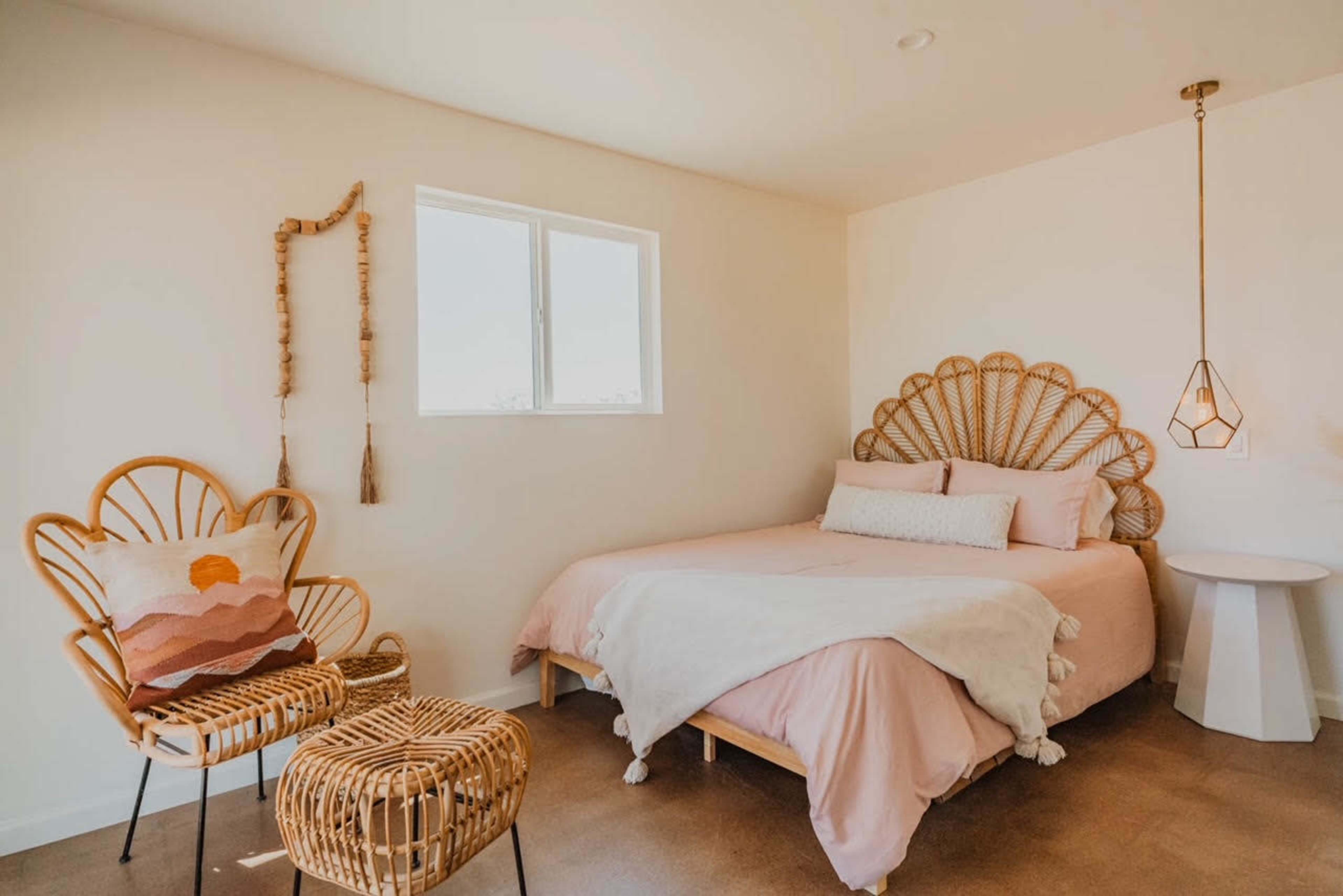 The room features a bed with a pink coverlet, a rattan headboard, and matching wicker chairs, set against a light-colored wall with a window.