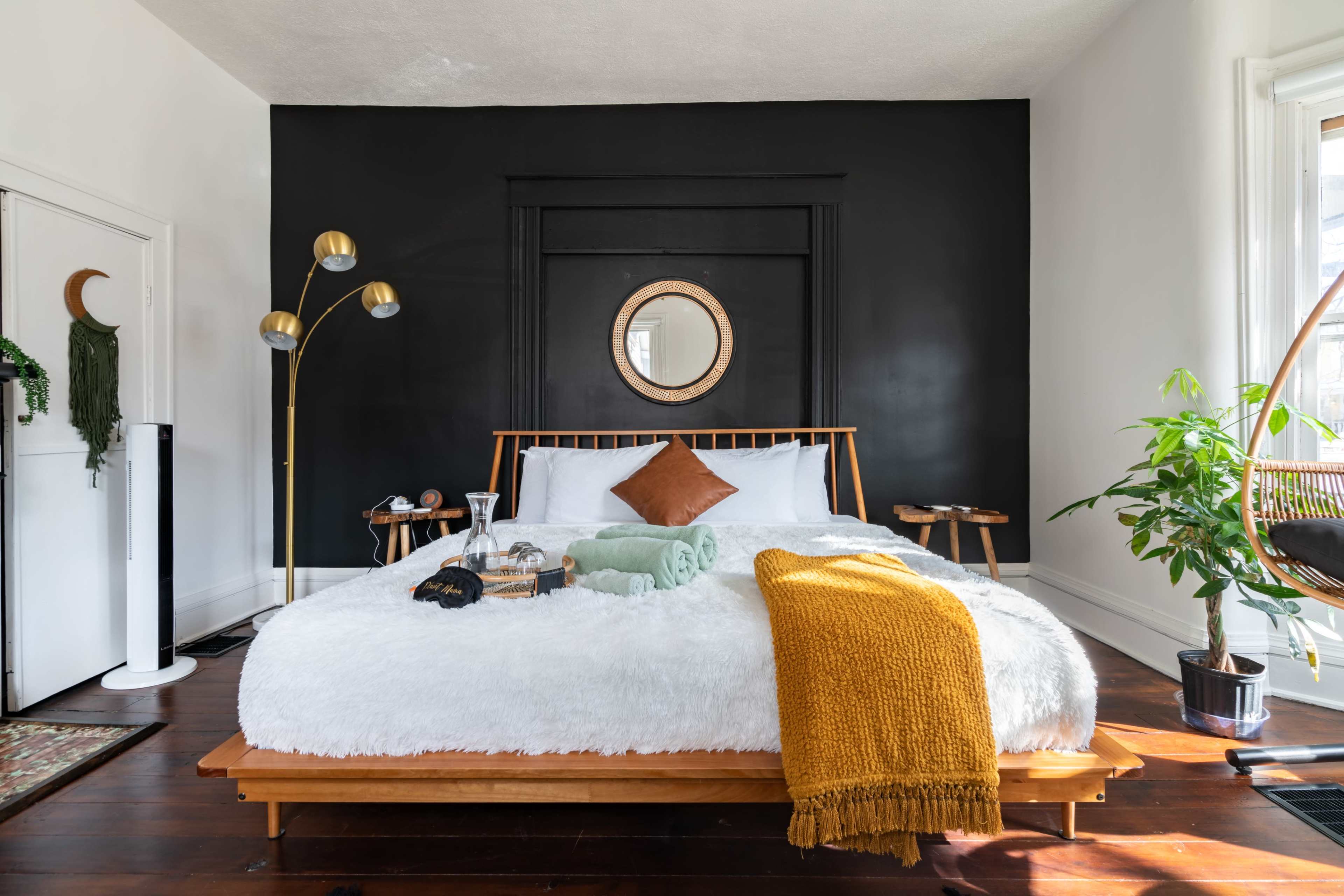 A cozy bedroom features a large fur-covered bed with a mustard blanket, flanked by side tables and a floor lamp against a dark accent wall.