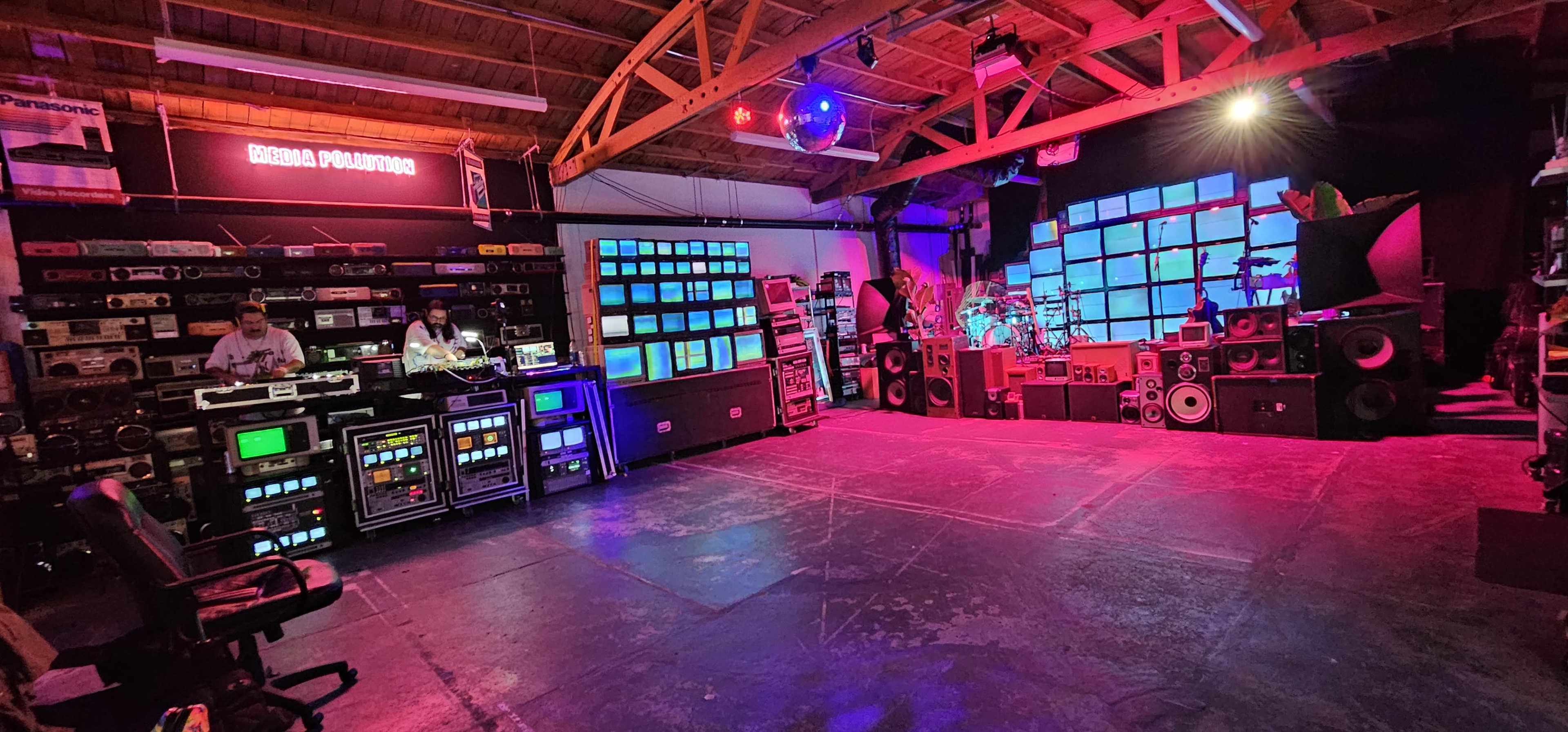 The image shows a spacious room filled with various audio and visual equipment, featuring multiple screens and speakers along the walls, and two individuals working at a mixing console.