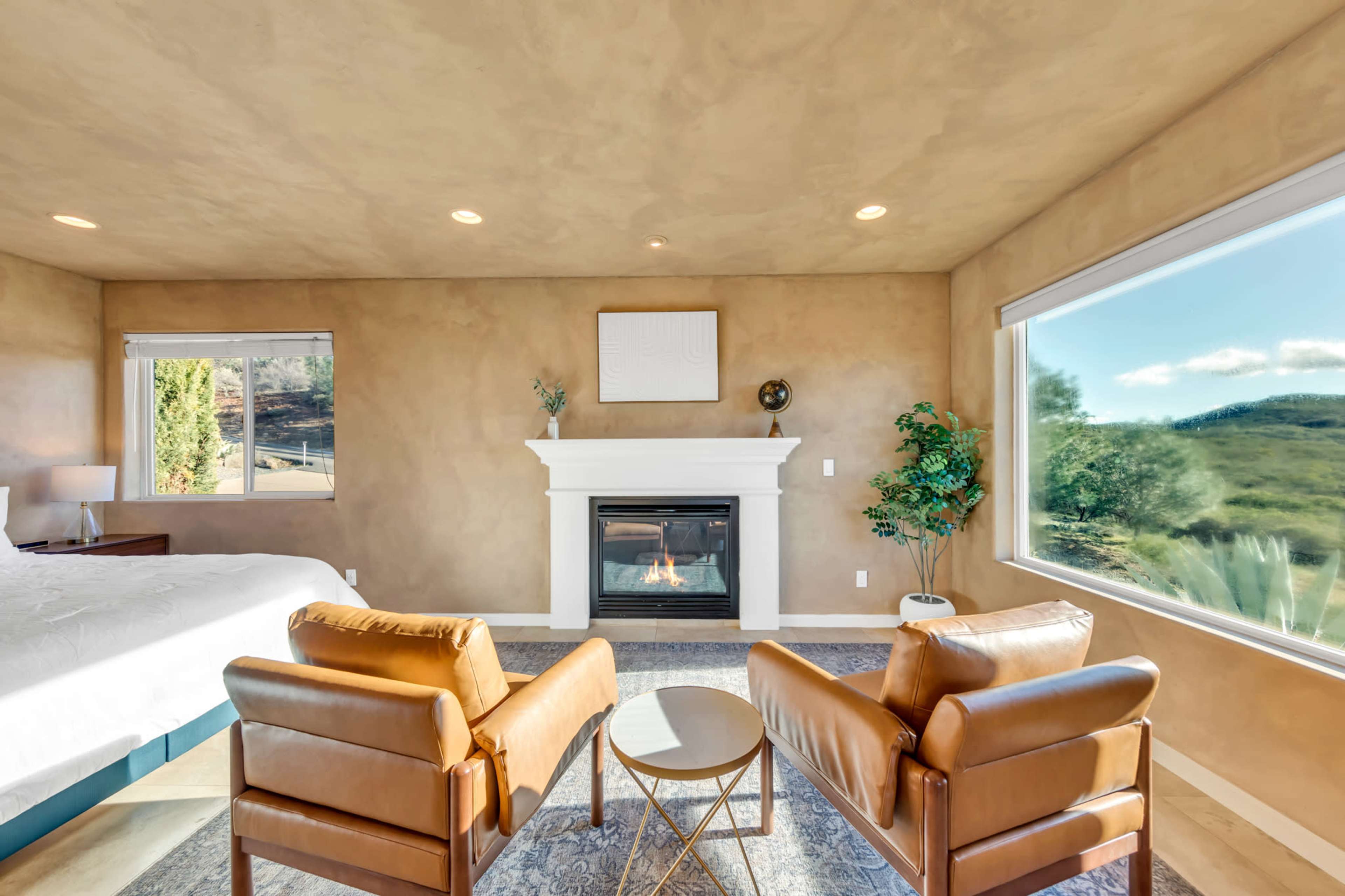A cozy room features two leather chairs facing a fireplace, with large windows showcasing a view of green hills outside.