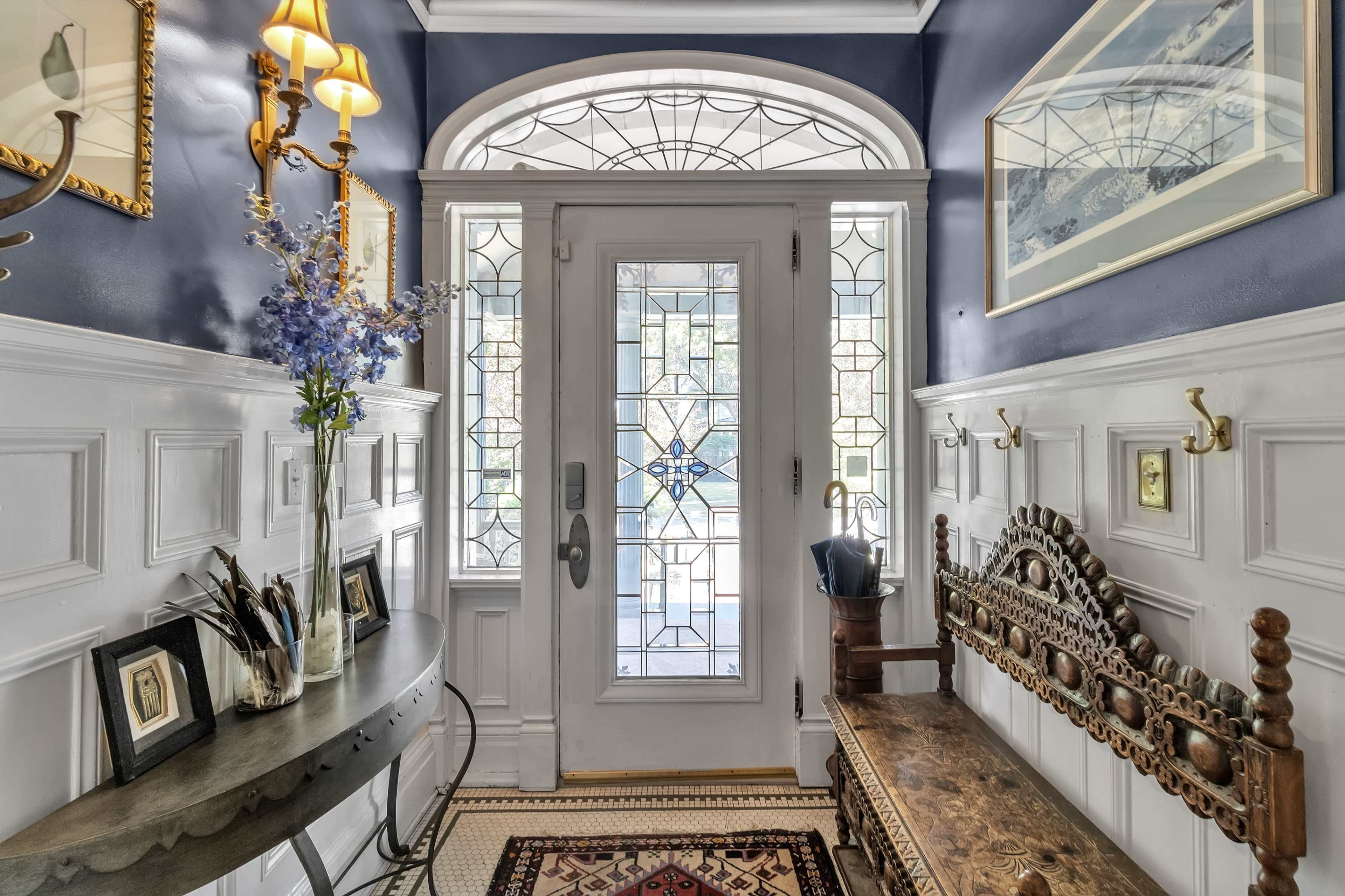The image shows an entrance hallway featuring a decorative bench, a small table with framed photos, and a stained glass door surrounded by blue walls.