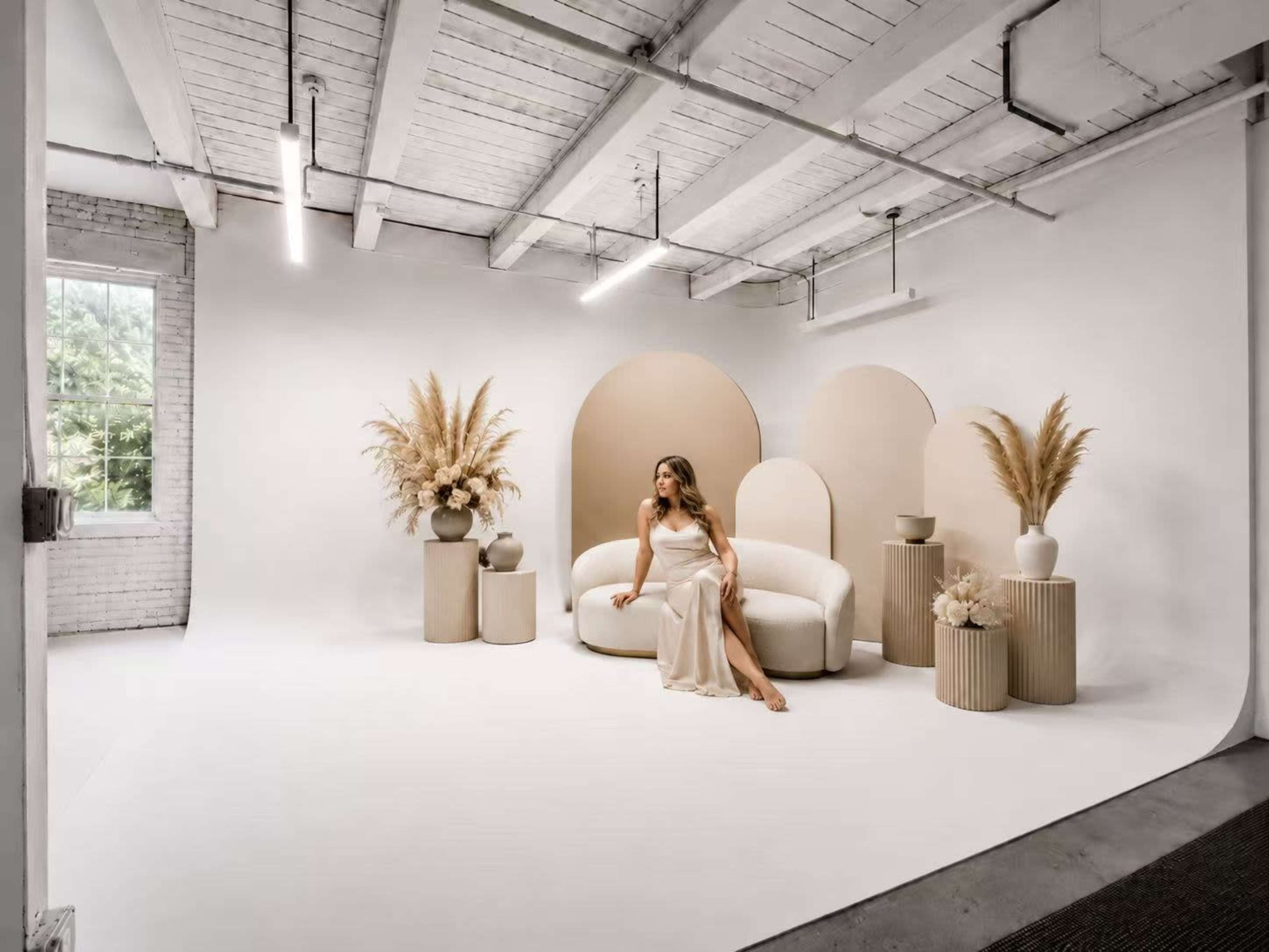A woman sits on a curved white sofa in a minimalist studio featuring neutral-colored decor and tall pampas grass arrangements.