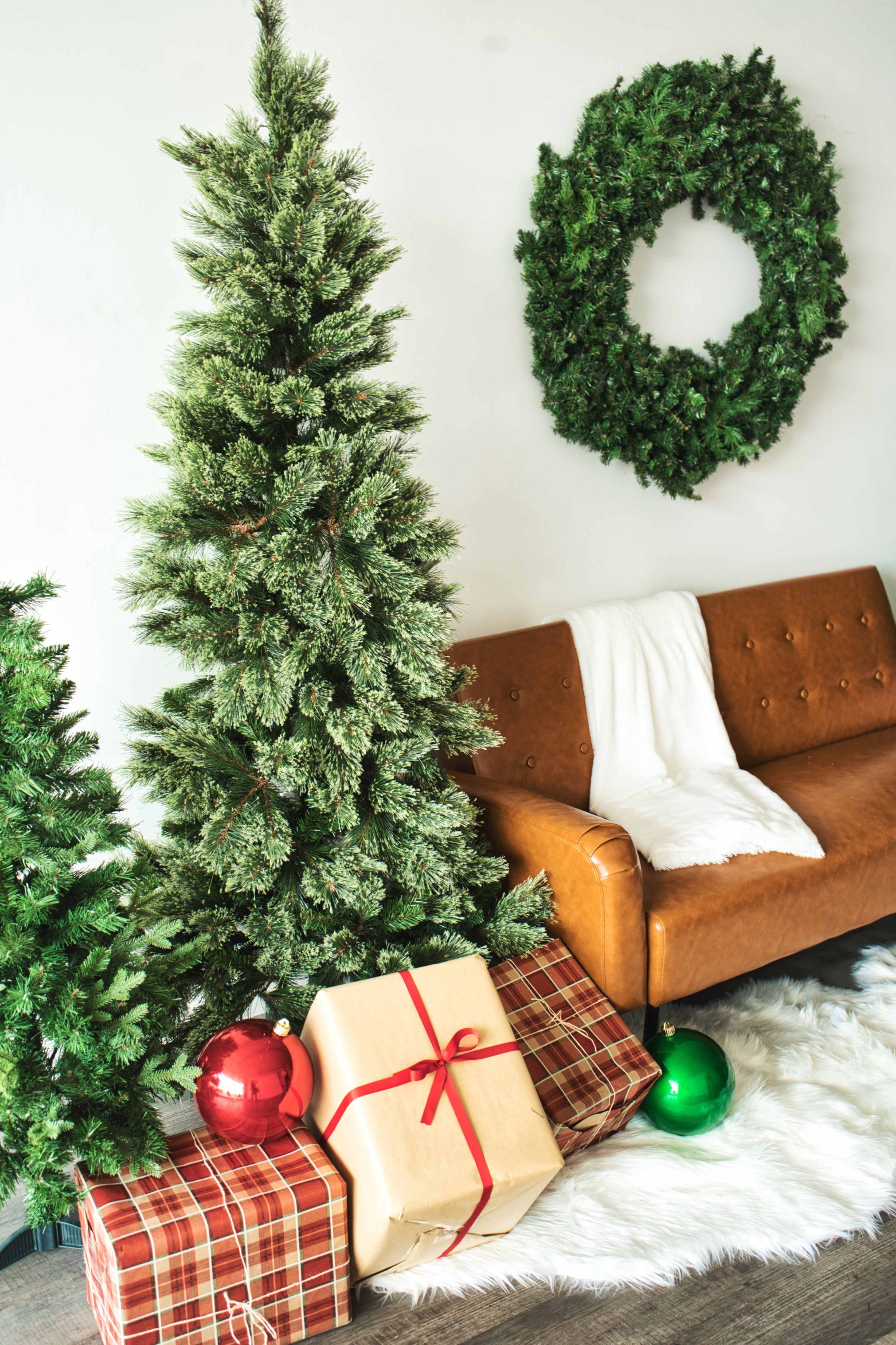 A decorated Christmas tree stands next to a cozy brown sofa, surrounded by wrapped gifts and holiday ornaments against a backdrop of a green wreath on the wall.