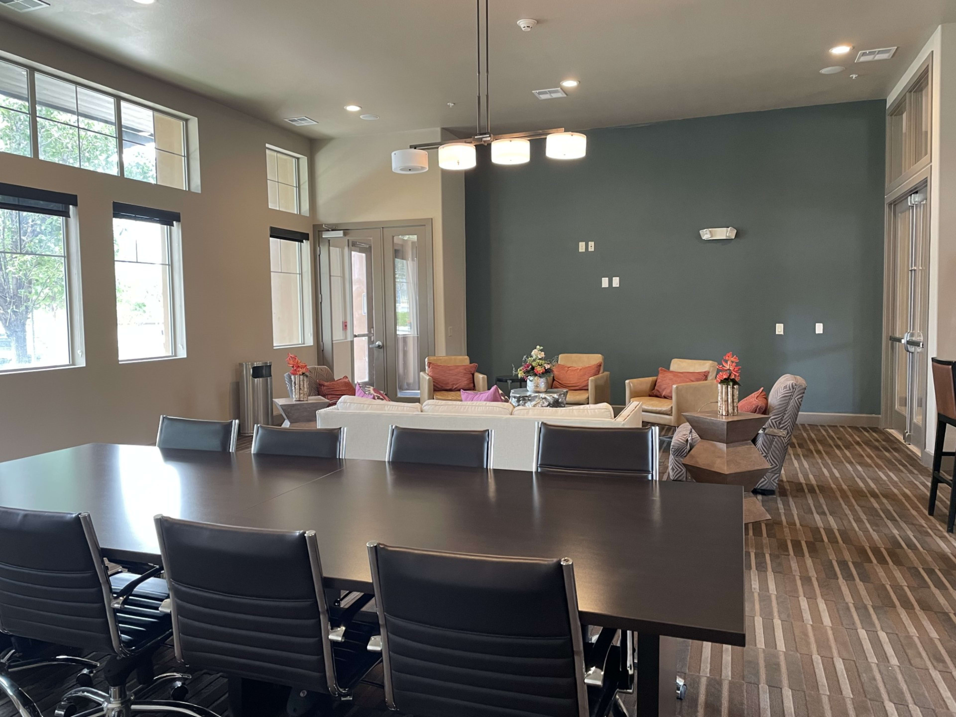 The image shows a modern lounge area with a large table and several seating arrangements, including couches and chairs, illuminated by overhead lighting.