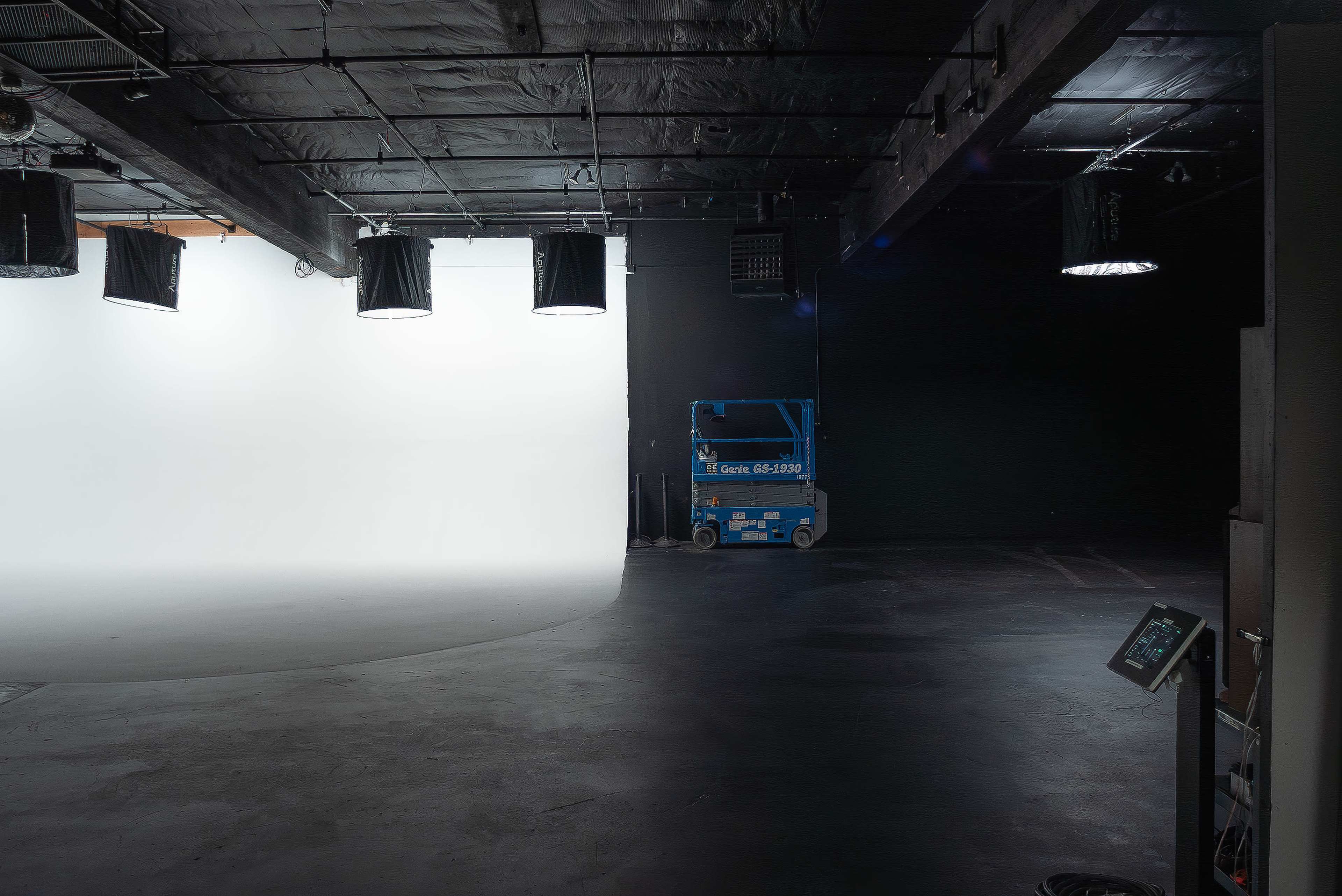 The image shows a large film studio with a white backdrop on one side and dark walls on the other, featuring lighting rigs and a scissor lift positioned in the center.