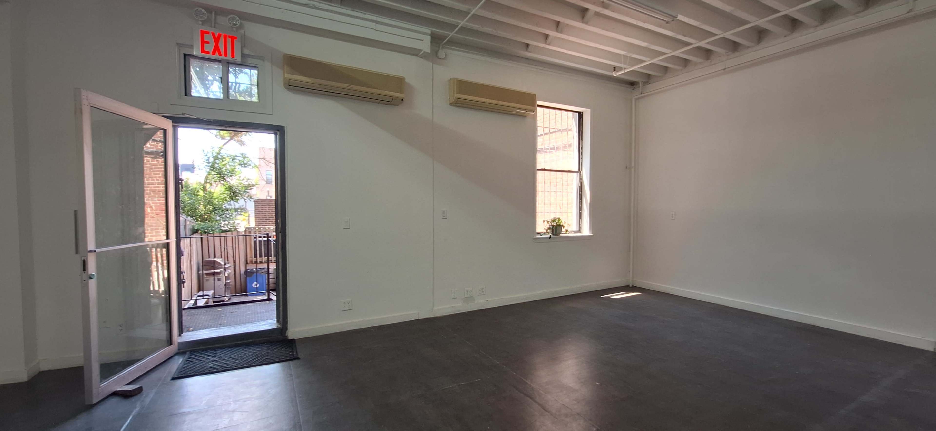 The image shows an empty room with hardwood floors, two air conditioning units on the walls, and an open door leading to an outdoor area.