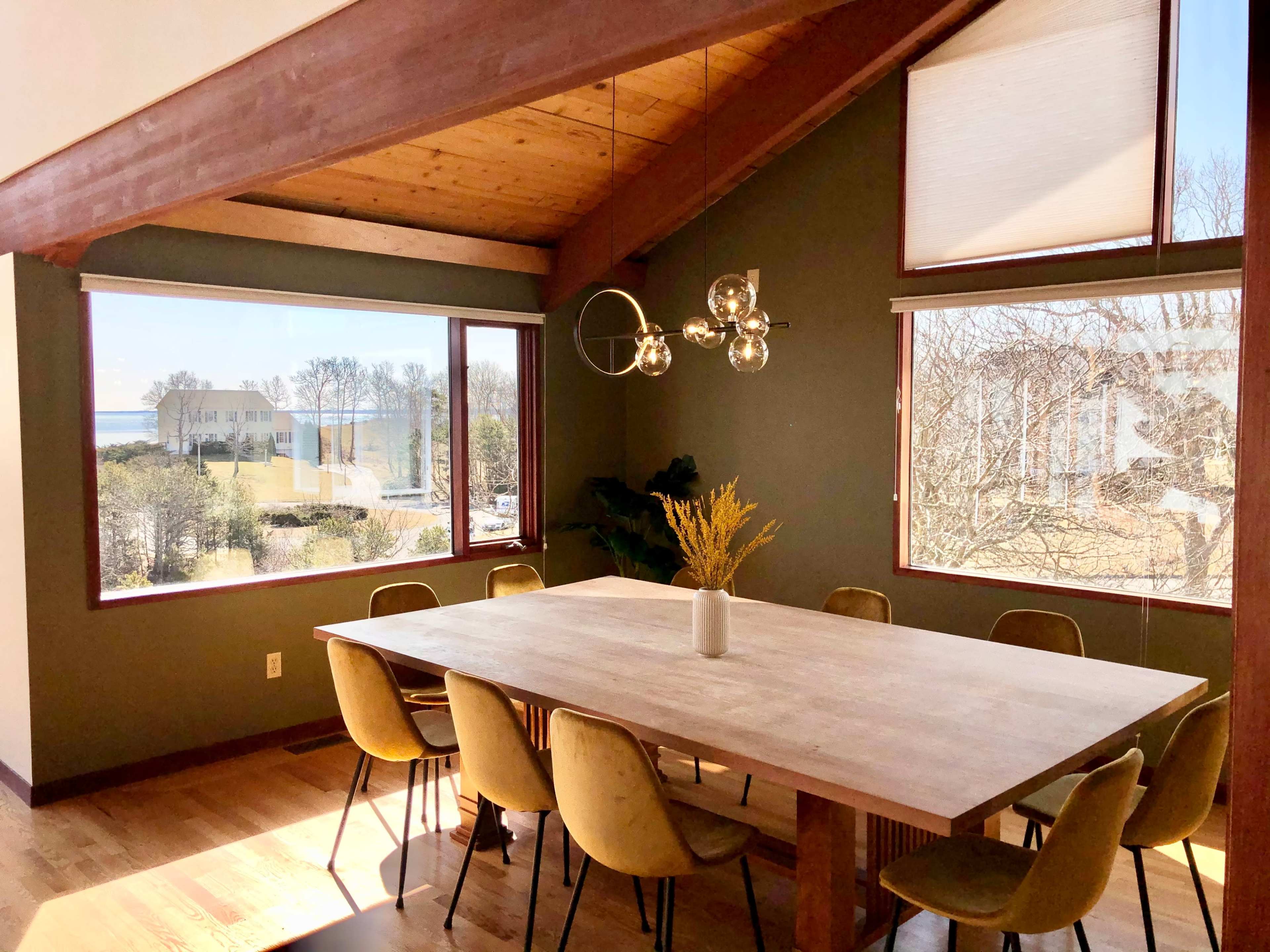 A spacious dining area features a large wooden table surrounded by upholstered chairs, with large windows offering a view of the landscape outside.