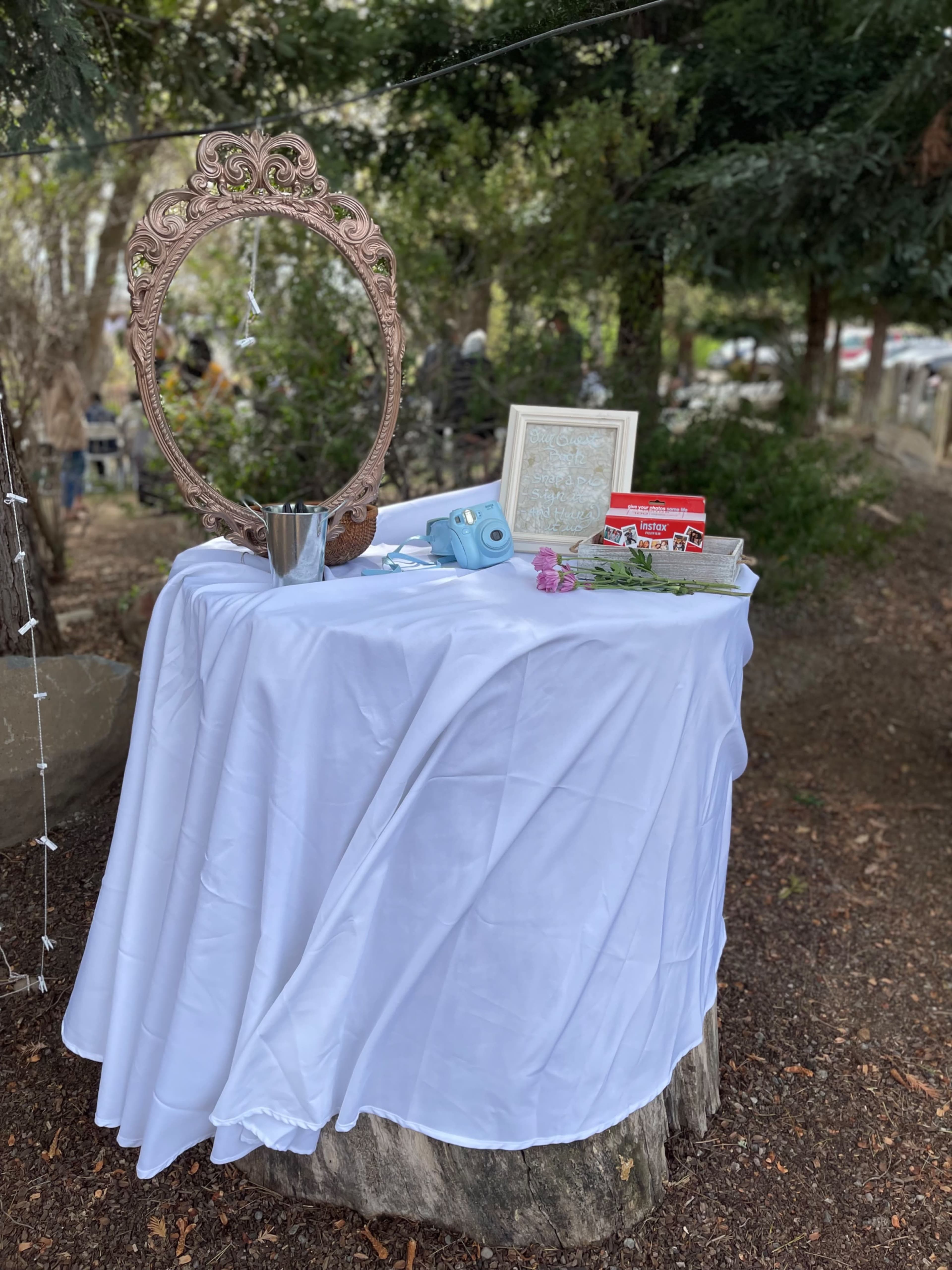 A white tablecloth drapes over a wooden stump adorned with a vintage mirror, a picture frame, a camera, a small vase with flowers, and decorative items in a wooded area.