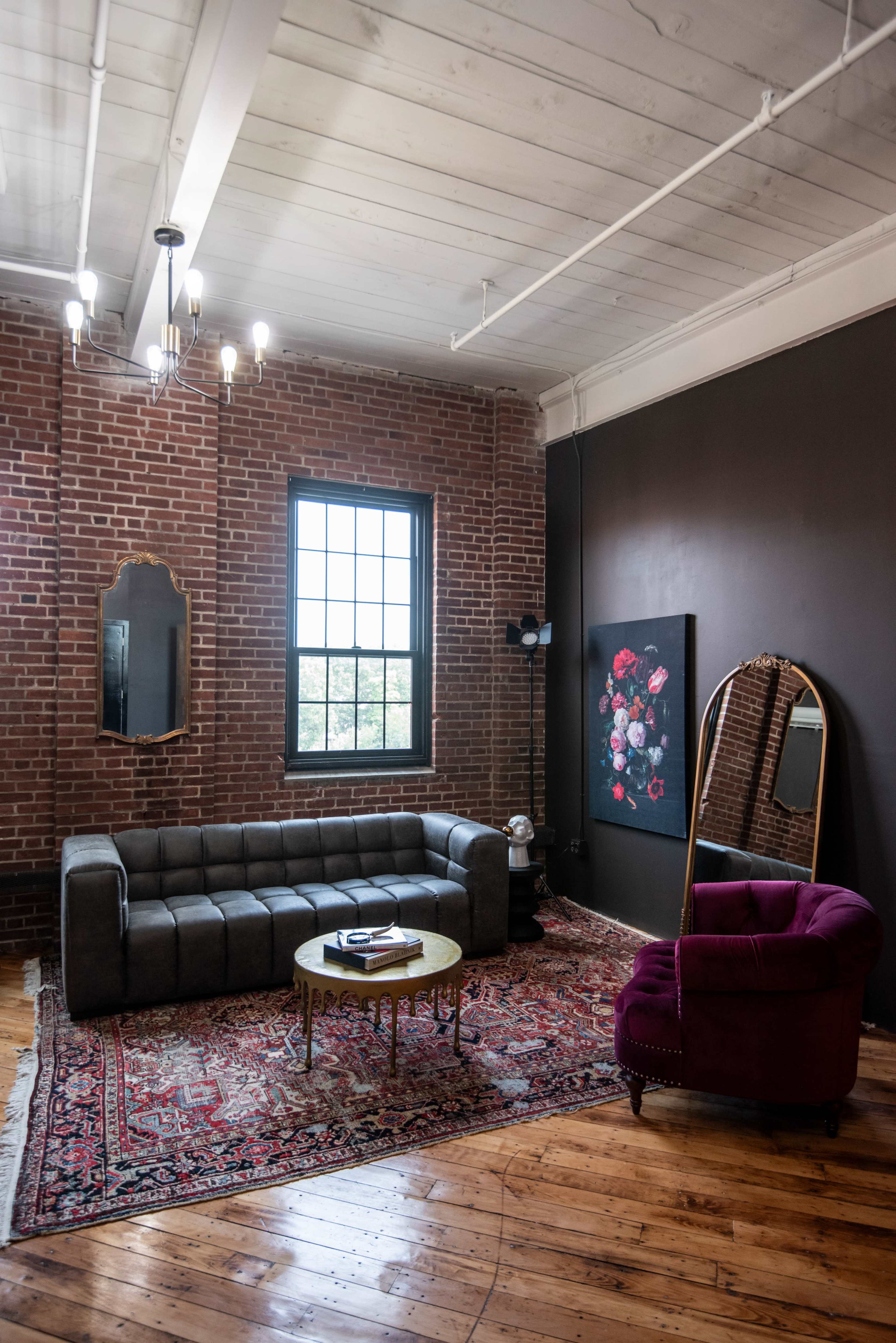 A living room with a brick wall, a large window, a gray sofa, a circular coffee table, a purple armchair, and a framed floral painting.
