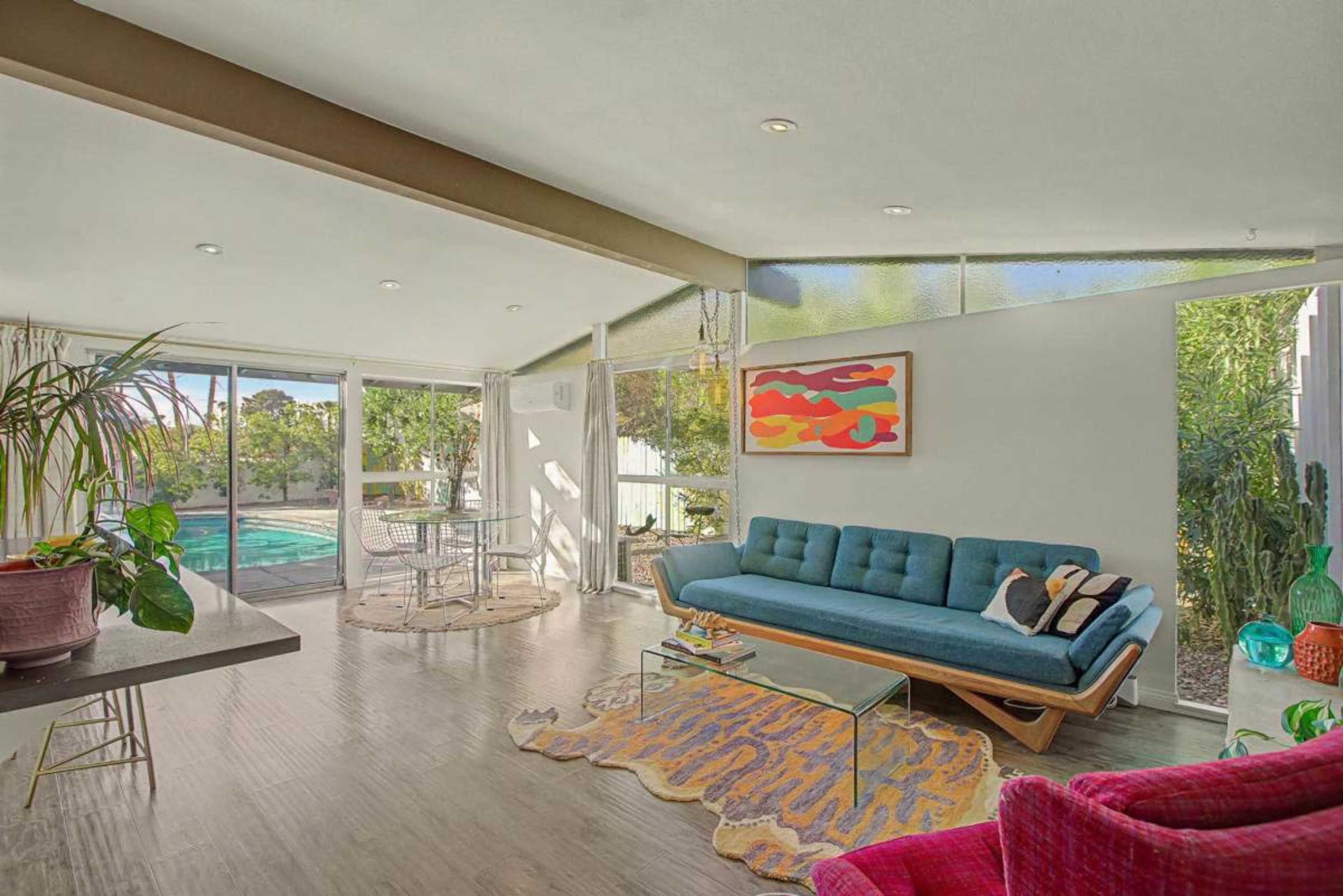 The image shows a modern, airy living room with a blue couch, a glass dining table, and large windows that overlook a pool and garden area.