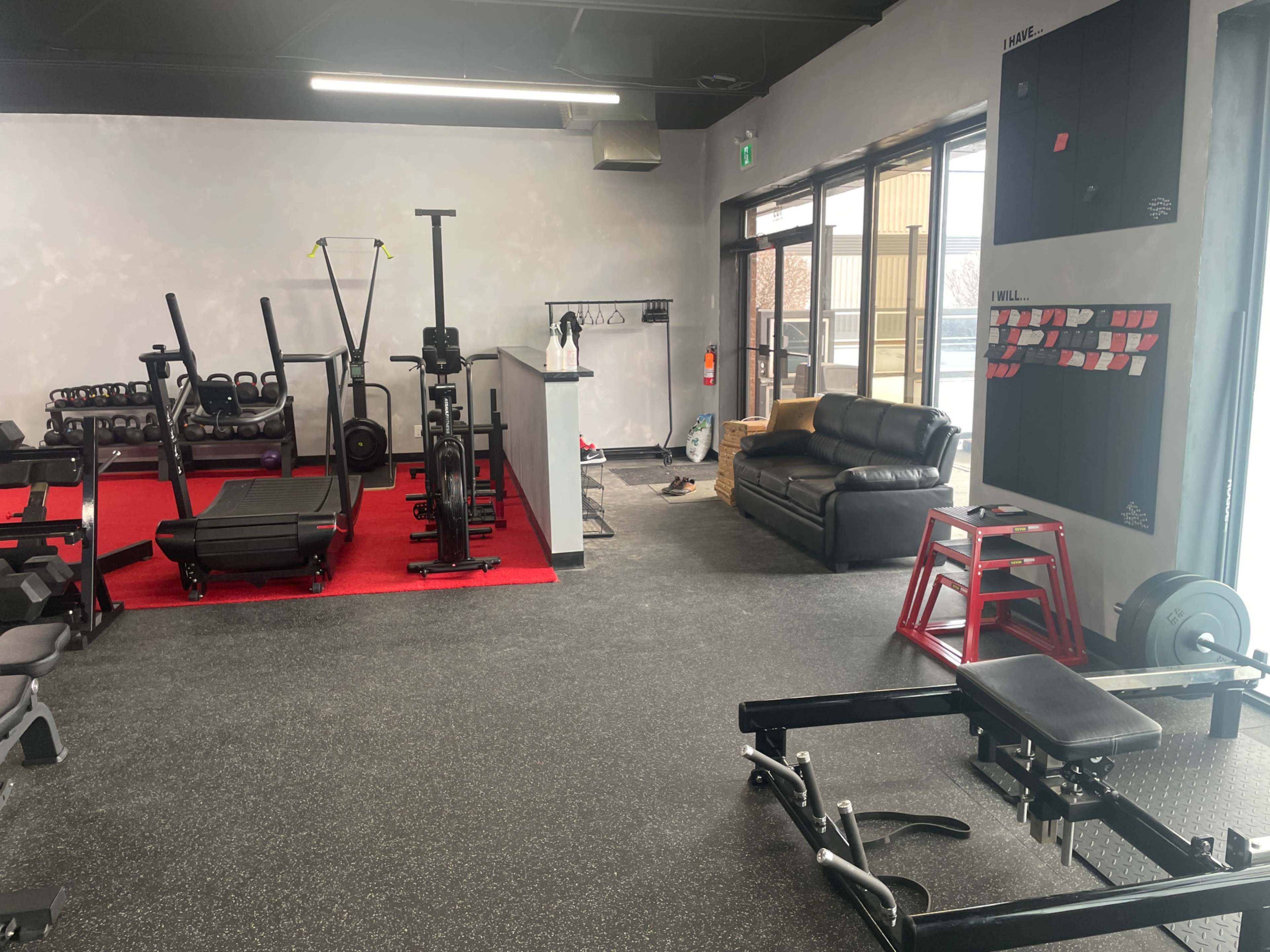 The gym interior features various fitness equipment, including weights, benches, and exercise machines, with a red carpet area and a seating space.