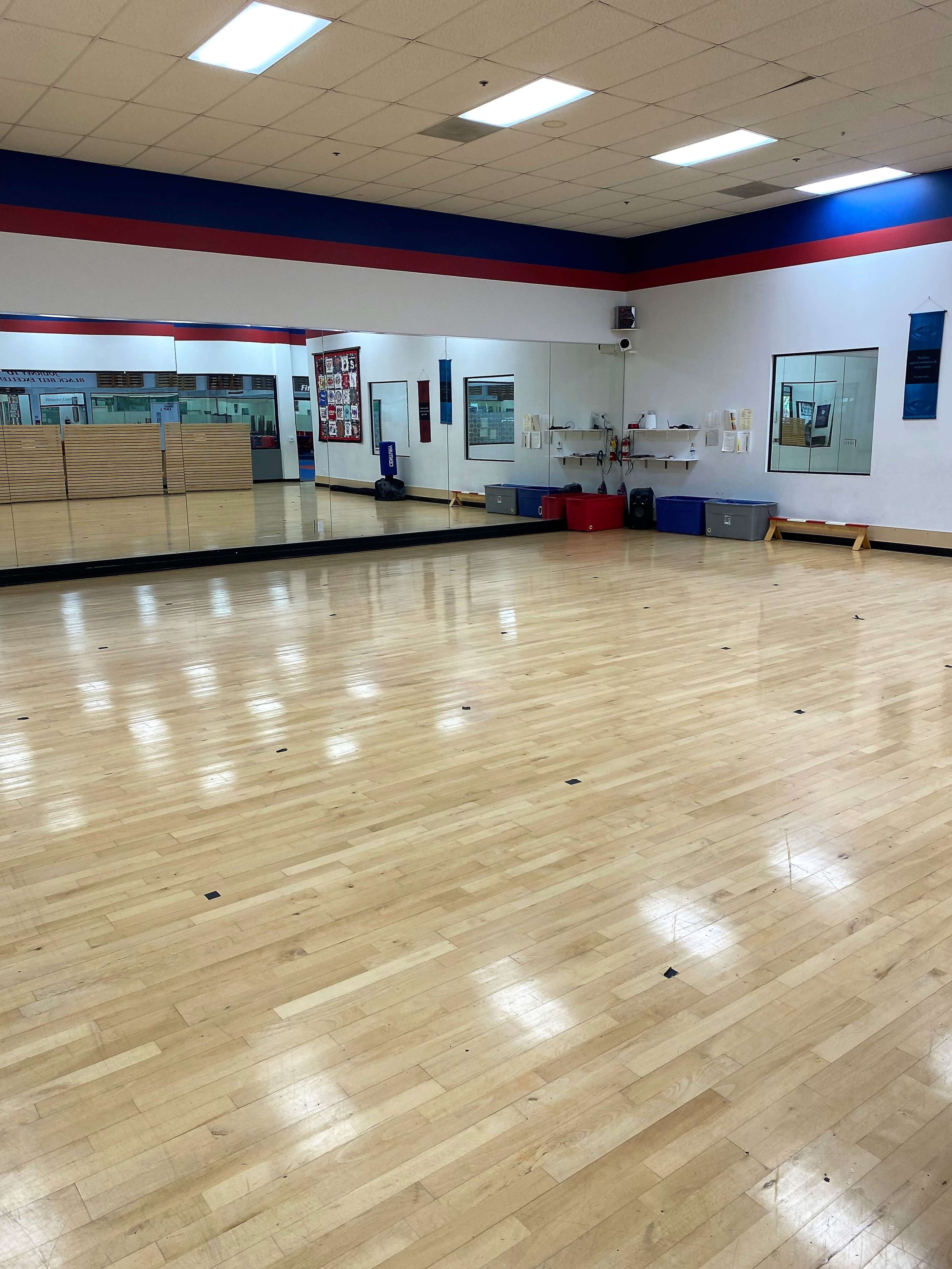 A large, empty dance studio with wooden flooring, mirrors on one wall, and storage boxes along the side.