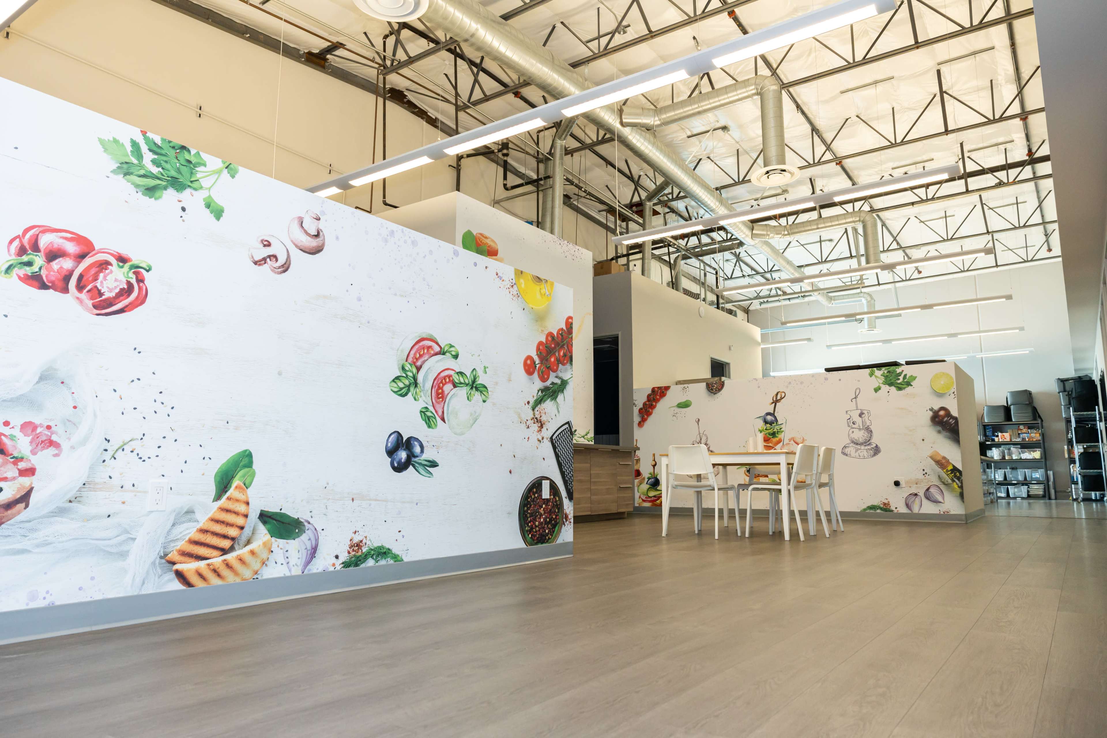 The image shows a spacious commercial kitchen area with decorated walls featuring food illustrations and a simple dining table with chairs.
