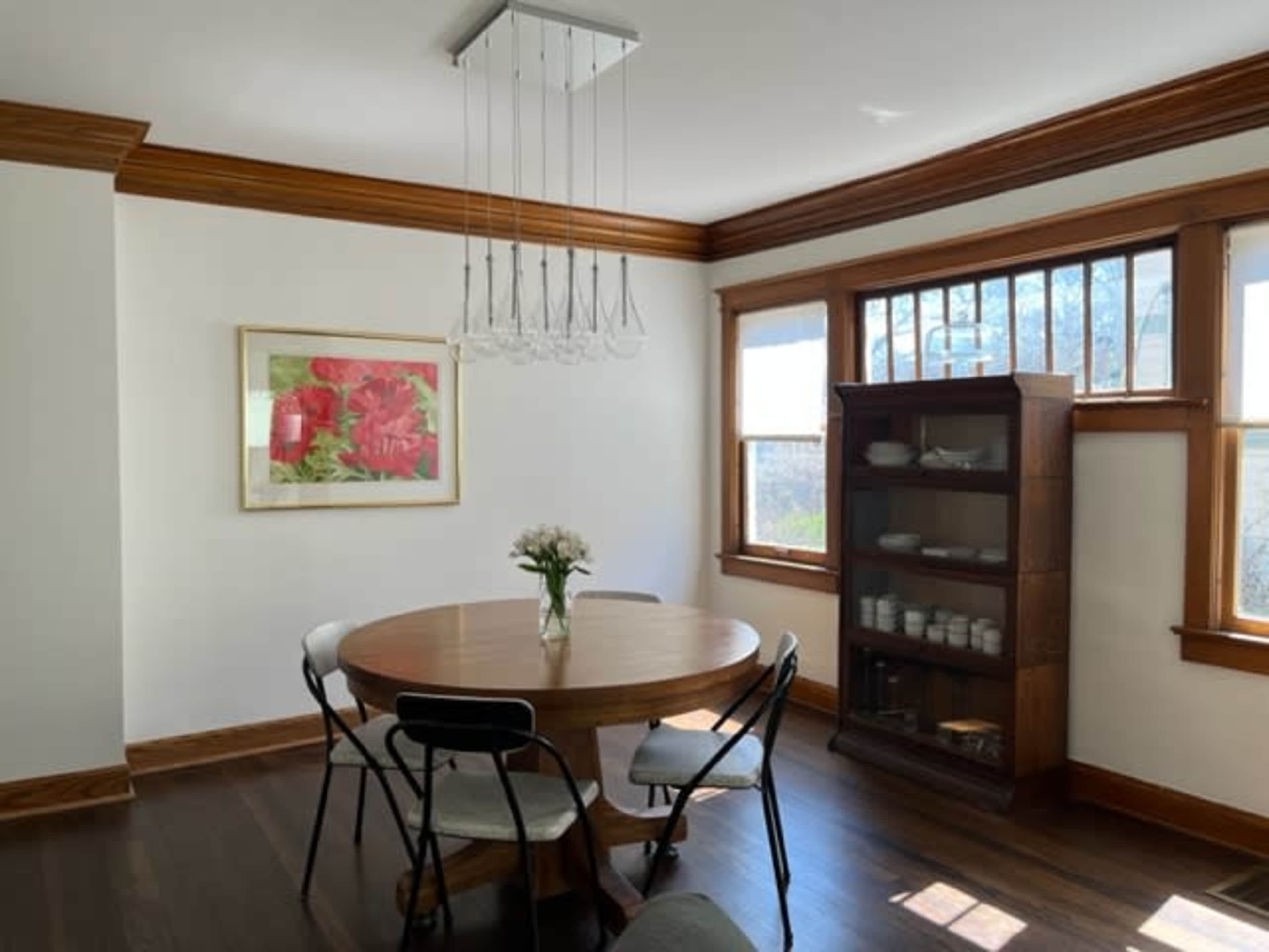 A round wooden dining table with four black chairs is set in a well-lit room featuring large windows and a vintage wooden cabinet.