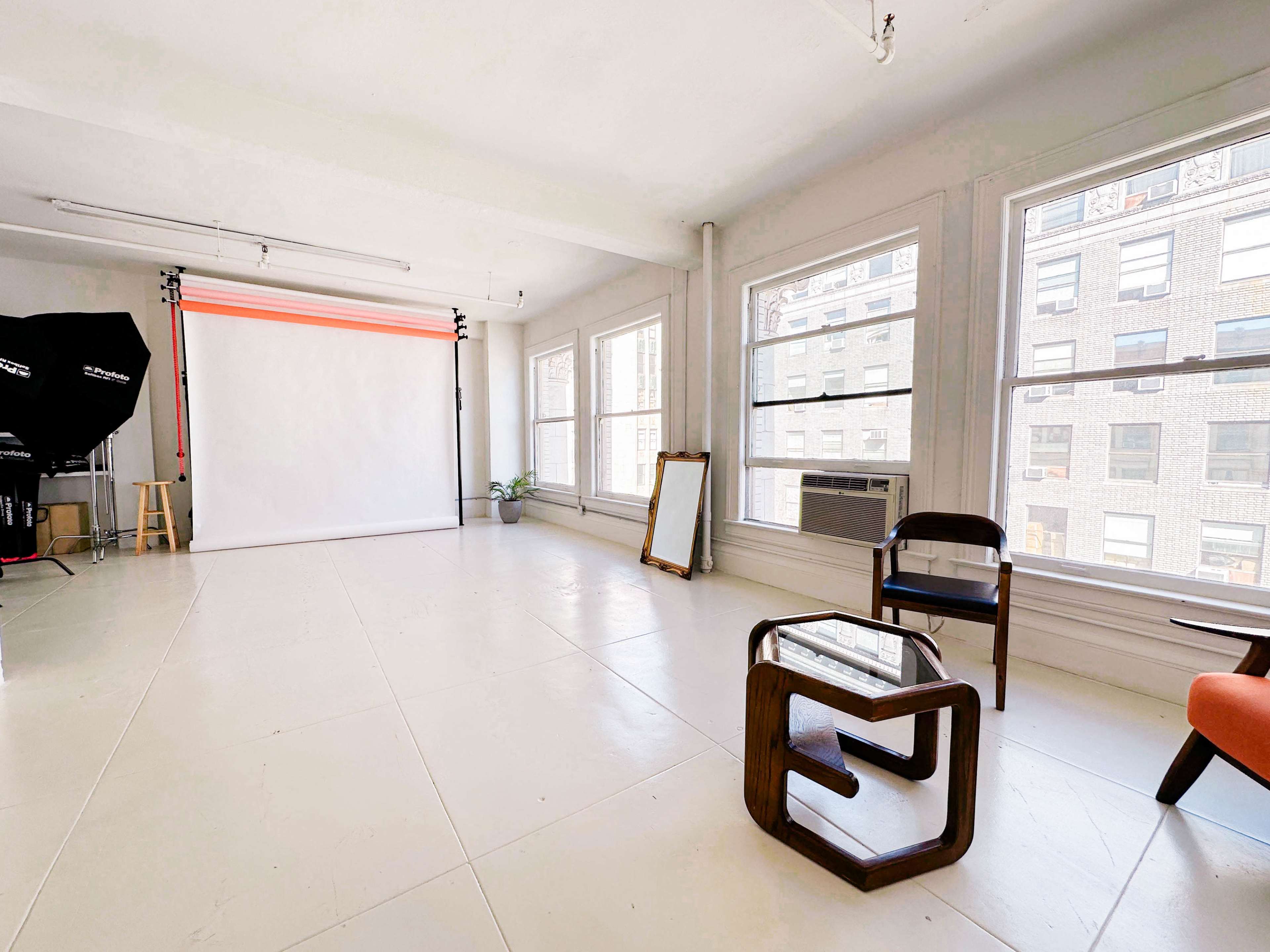 A bright, empty photography studio with a white backdrop, large windows, and minimal furniture including a chair and a coffee table.