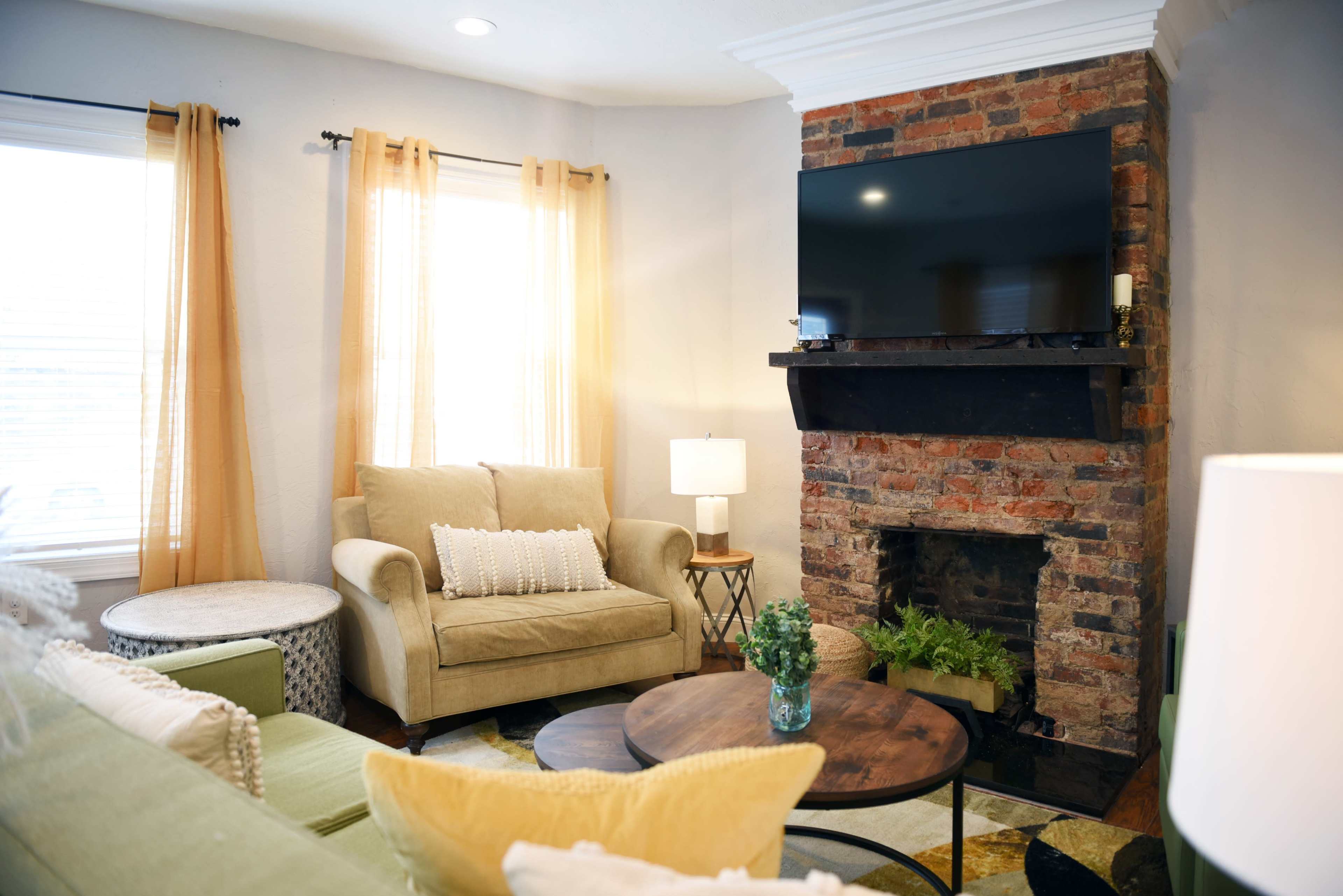 A cozy living room with a green sofa, a beige armchair, a brick fireplace, and a mounted television above it, illuminated by natural light from the windows.