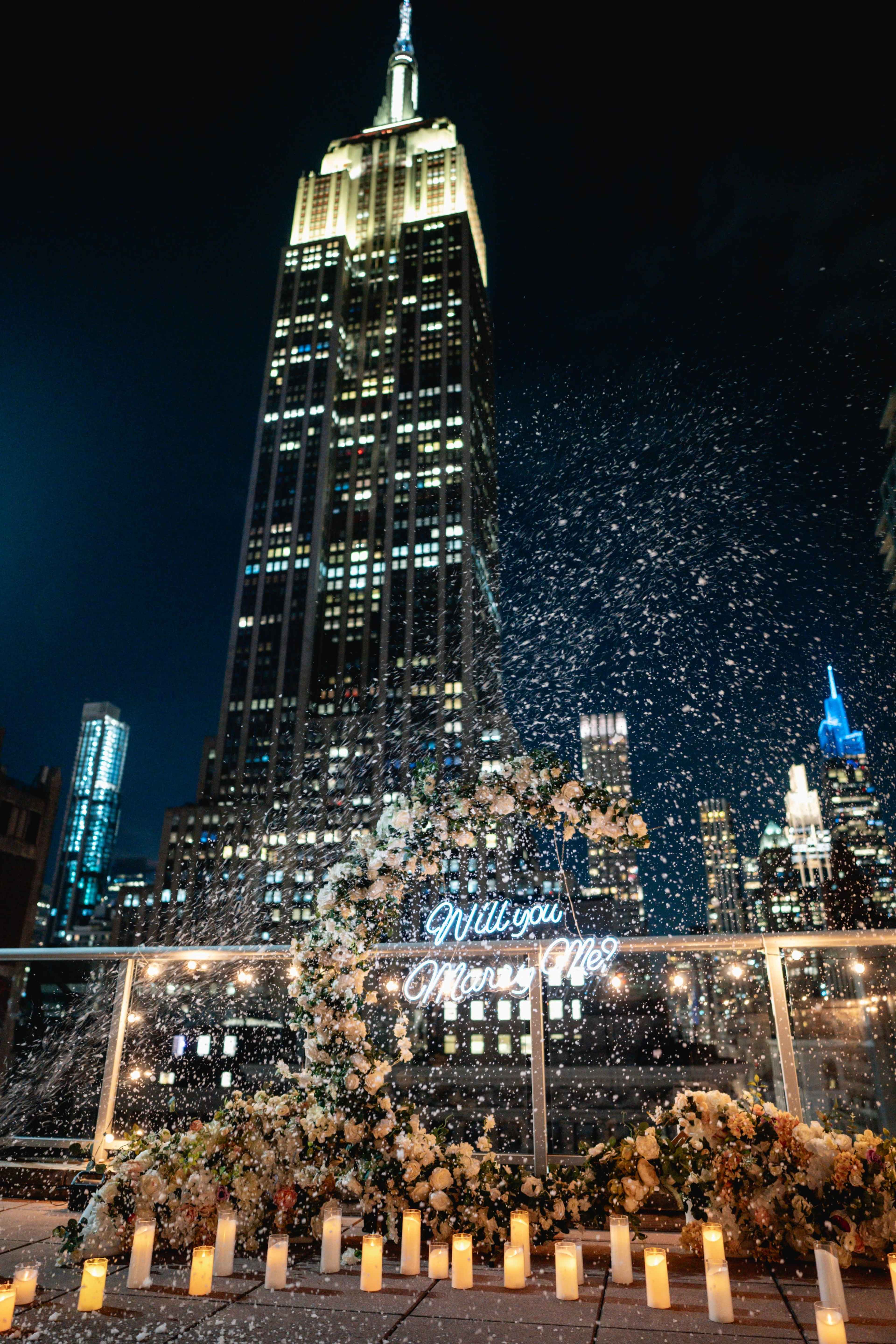 A romantic rooftop proposal scene features a glowing "Will you Marry Me" sign surrounded by flowers and candles, with the Empire State Building illuminated in the background.