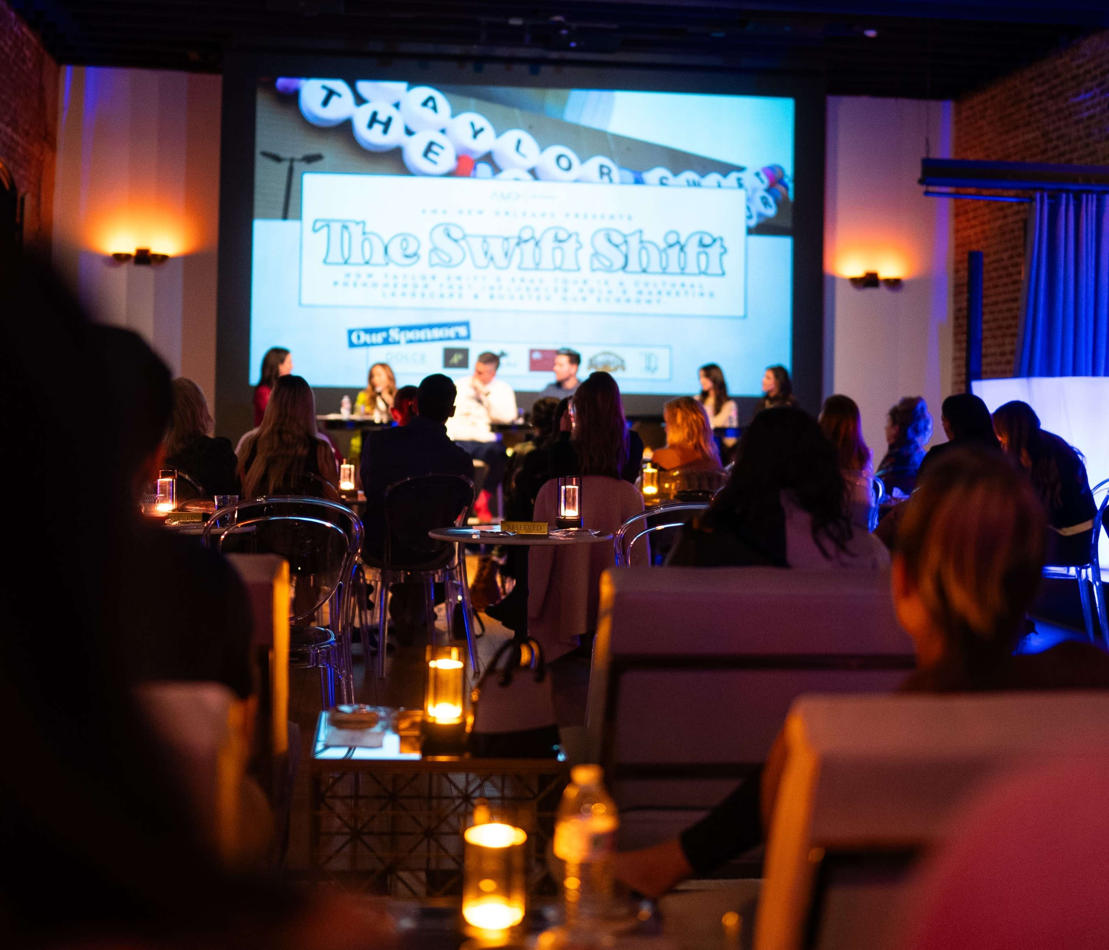 The scene depicts a panel discussion with several speakers seated at a table in front of an audience, illuminated by candles on tables, with a large screen displaying promotional graphics behind them.