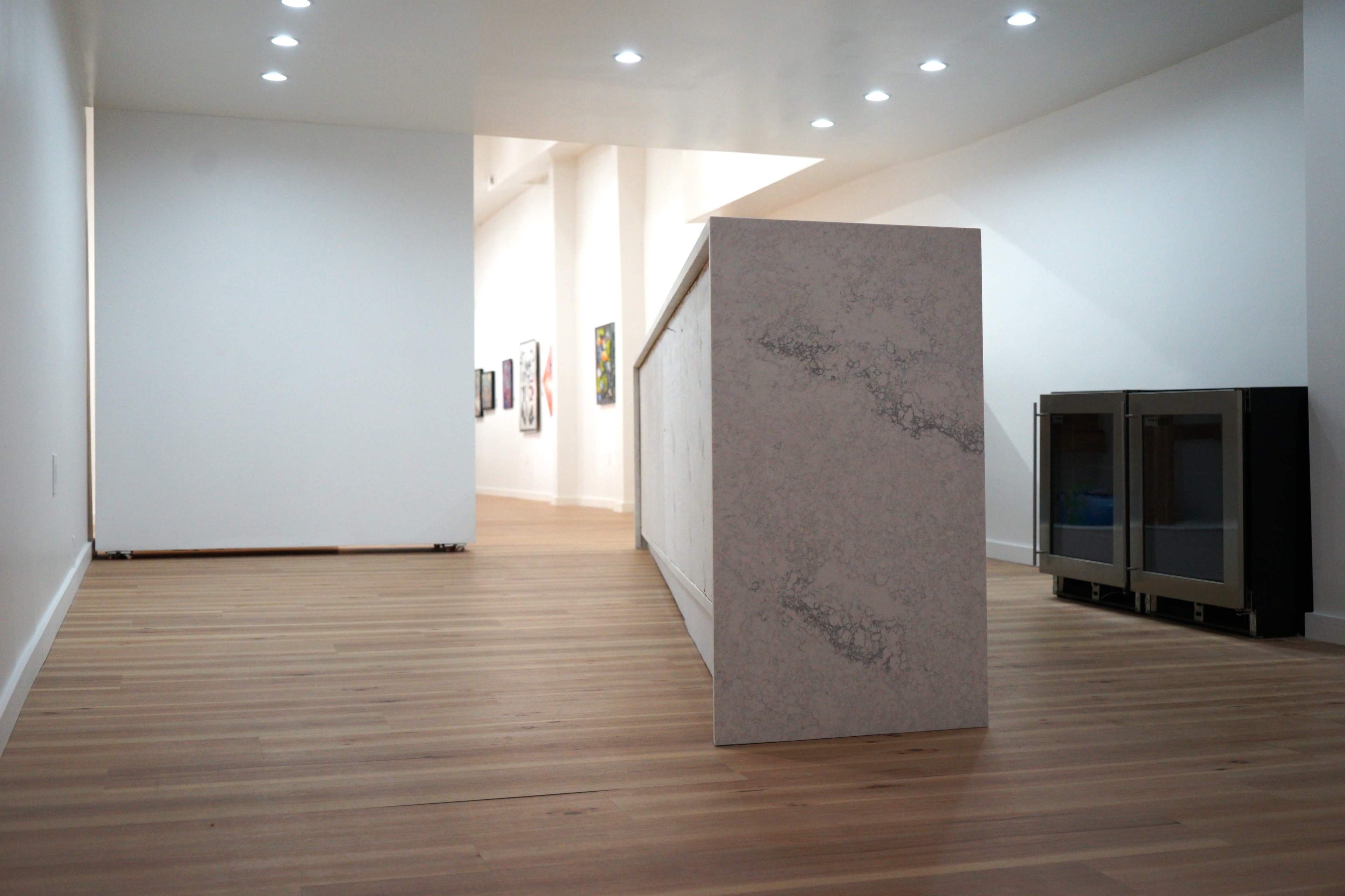 The image shows a modern interior space with light wood flooring, a large white counter or island, and a wall adorned with framed artwork in the background.