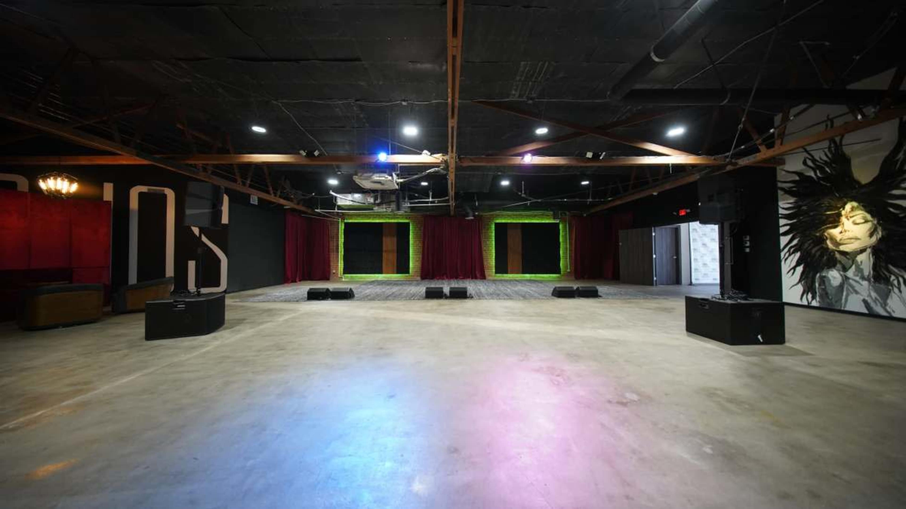 The image shows a spacious indoor venue with a polished concrete floor, a performance stage framed by curtains, and a mural on one wall.