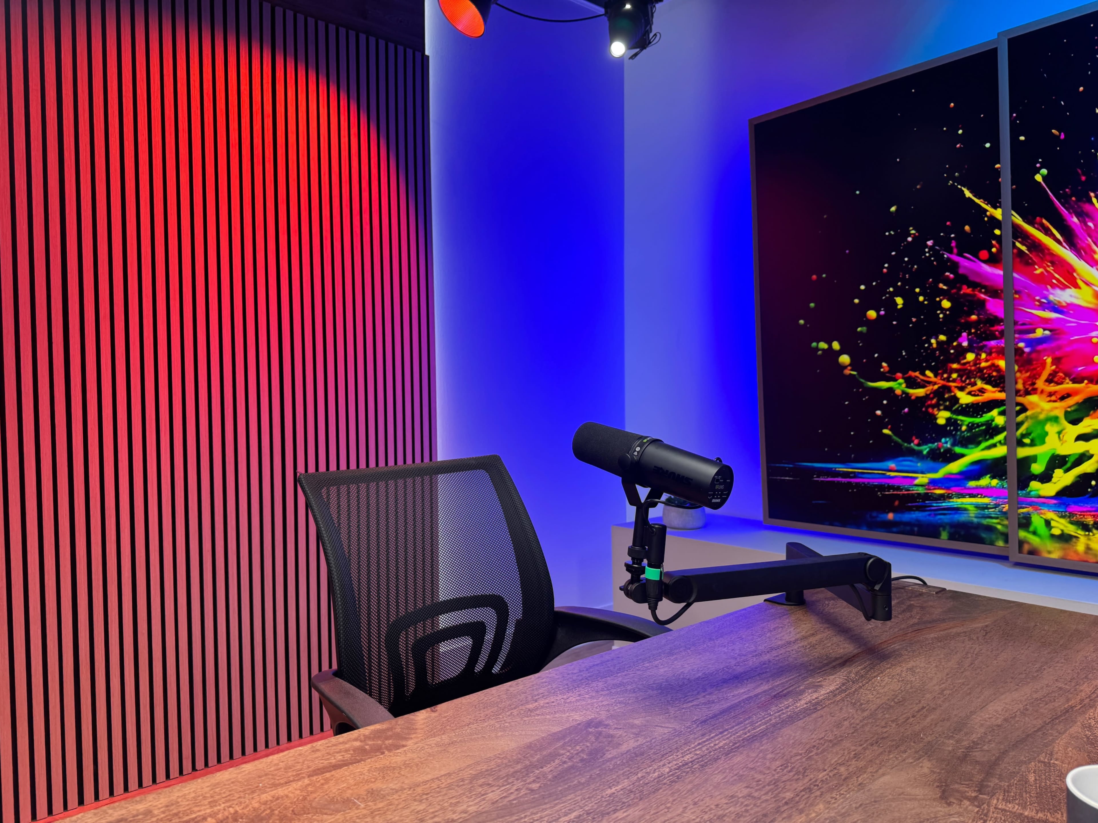 A well-lit recording studio setup features a closed-back chair and a microphone mounted on an arm, positioned beside a large, colorful display.