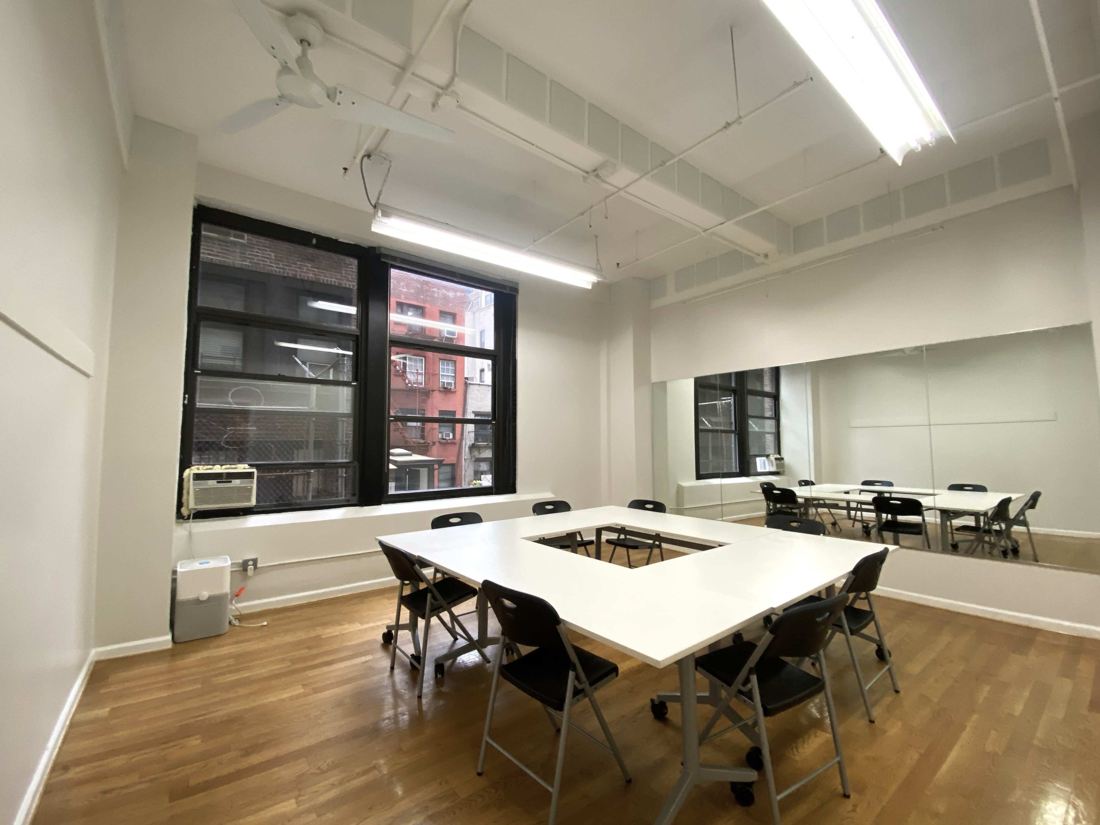 A meeting room features a square arrangement of tables and chairs, with large windows and a mirrored wall.