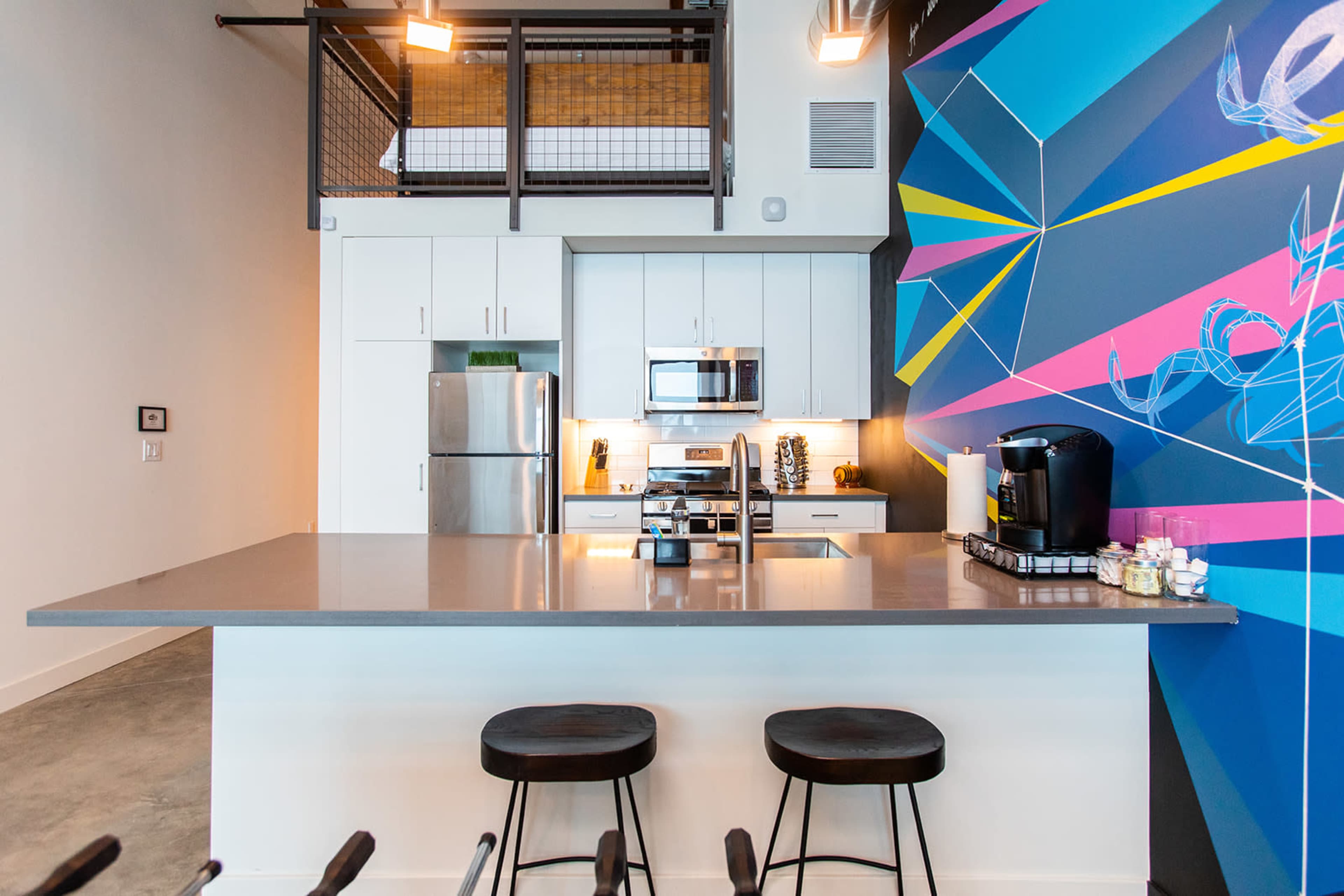 A modern kitchen with a colorful geometric wall mural, stainless steel appliances, and a sleek countertop with three black stools.