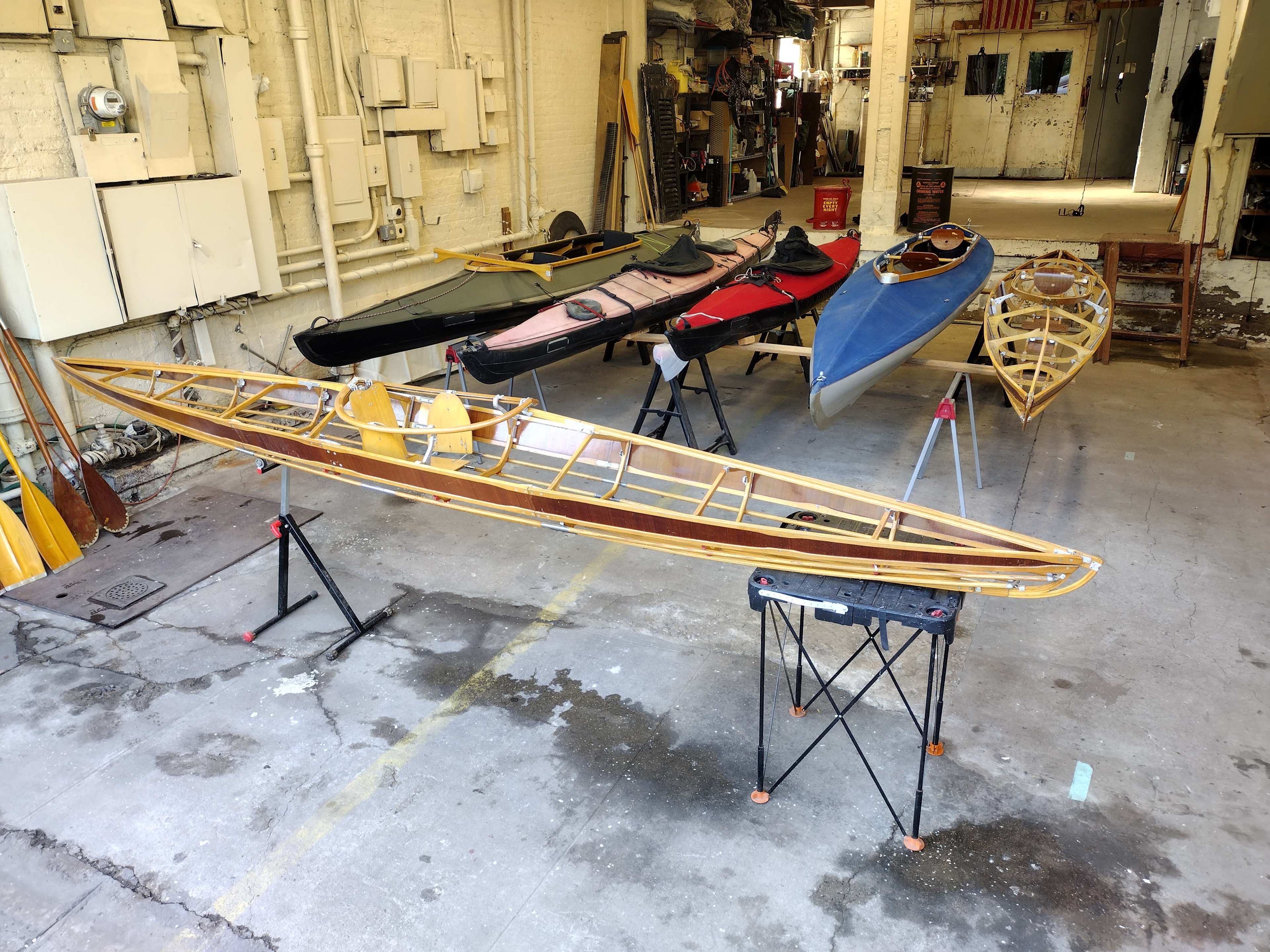 The image shows several kayaks displayed on stands inside a workshop, with tools and workstations in the background.