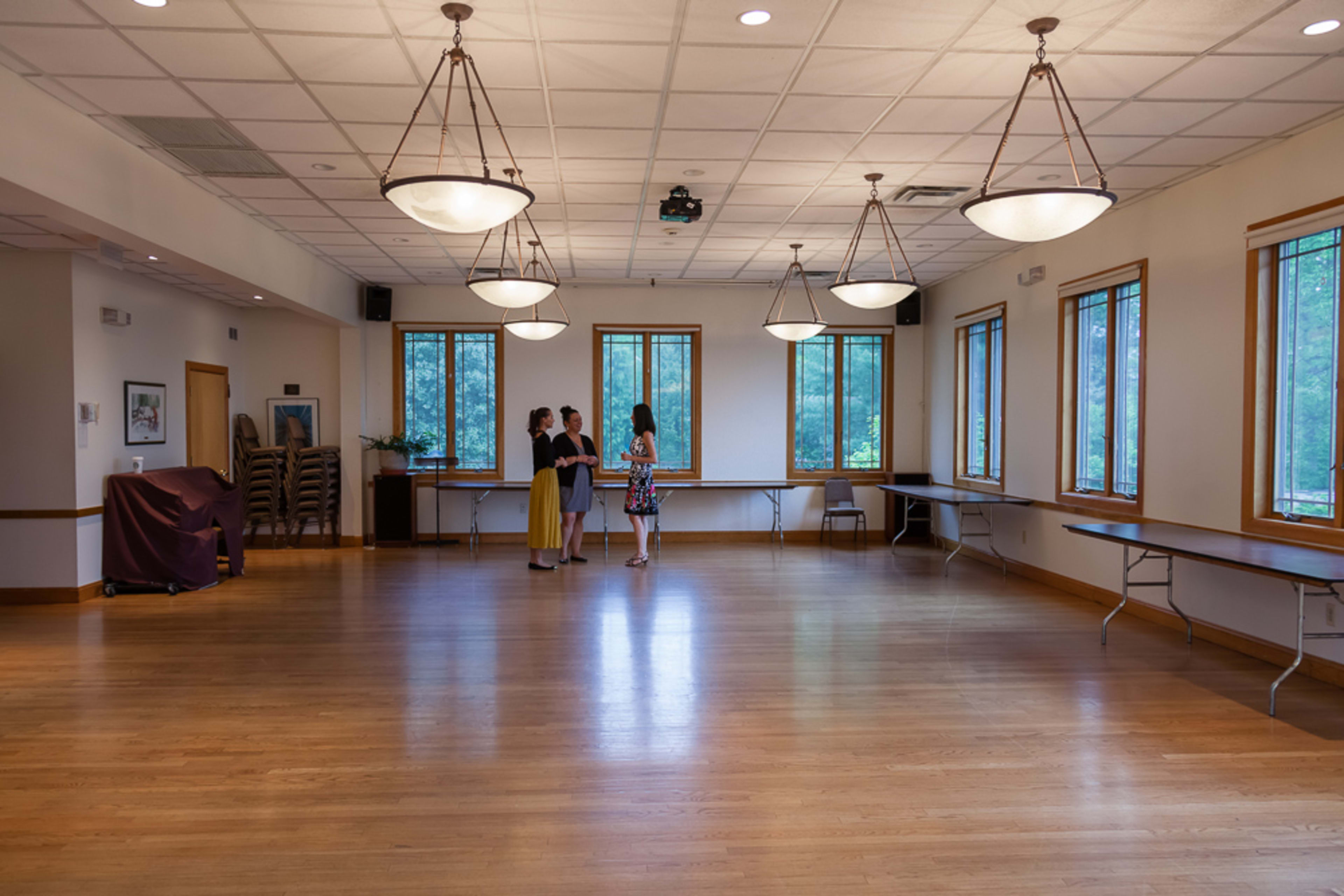 Three women stand together in a spacious, well-lit room with wooden floors and large windows, surrounded by folding chairs and tables.