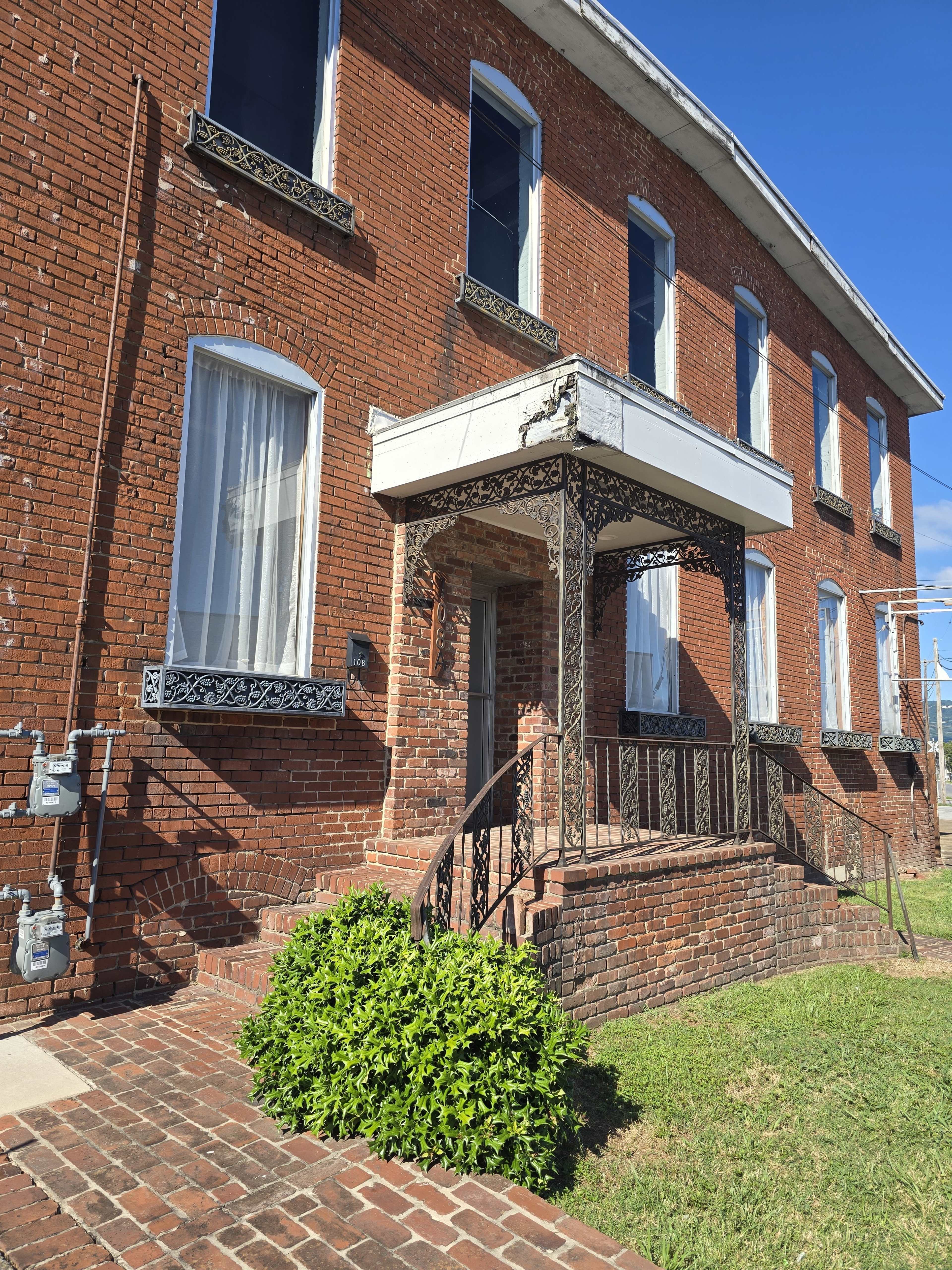 The image shows a brick building with a porch featuring intricate ironwork, large windows, and a well-maintained front yard.