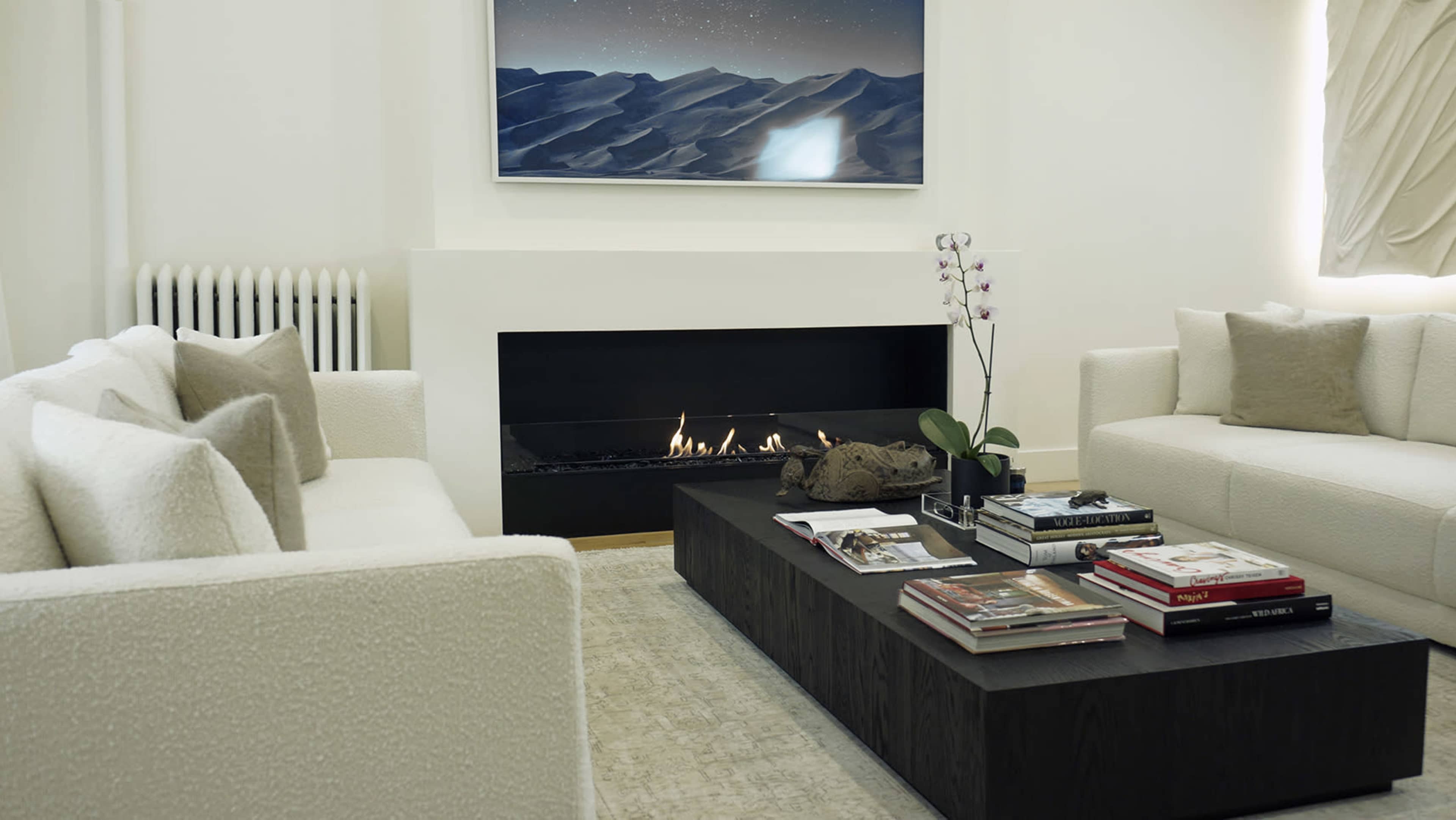A modern living room with two white sofas facing a central coffee table, a sleek fireplace underneath a wall-mounted artwork.
