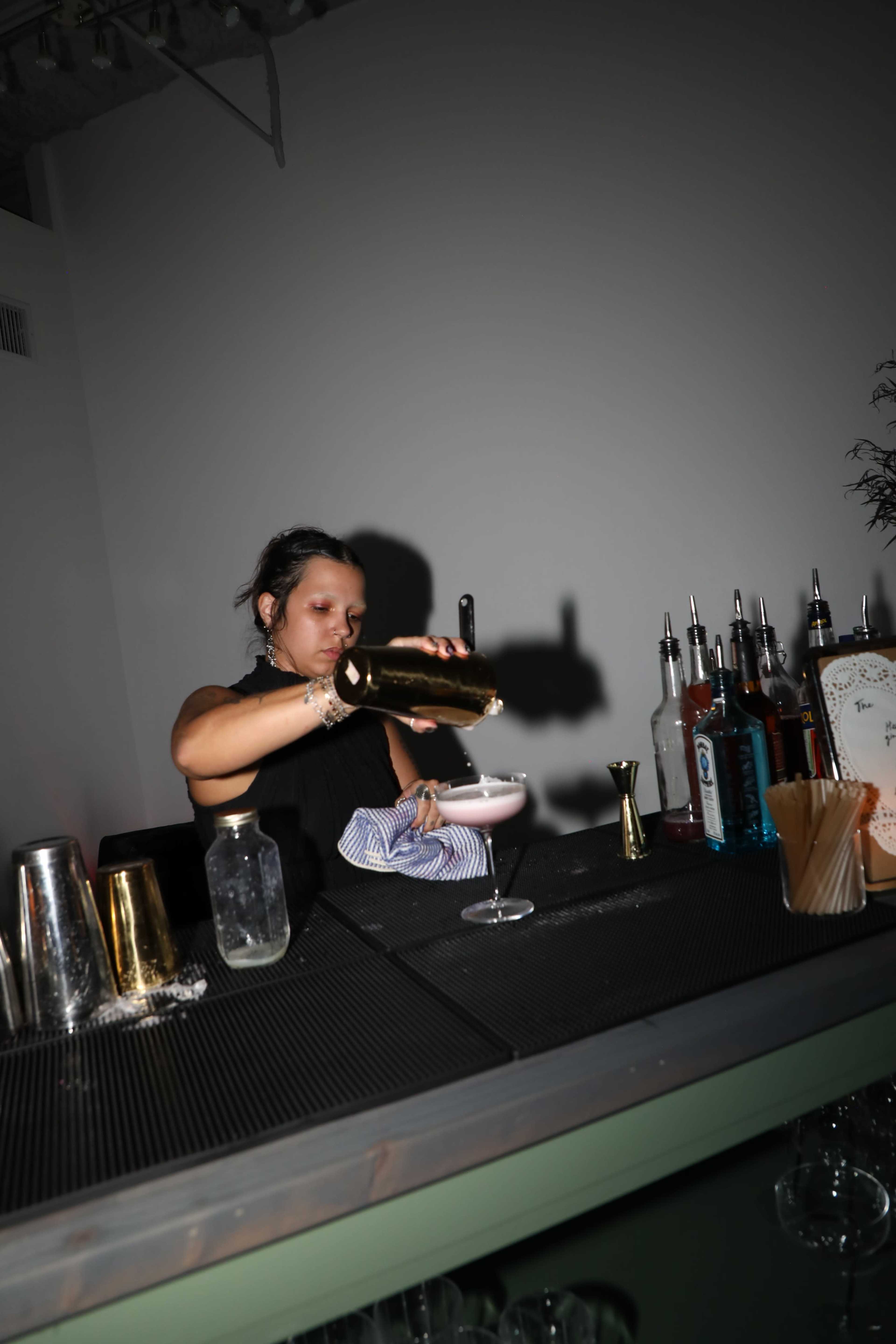 A bartender pours a cocktail from a shaker into a glass while working at a well-stocked bar.