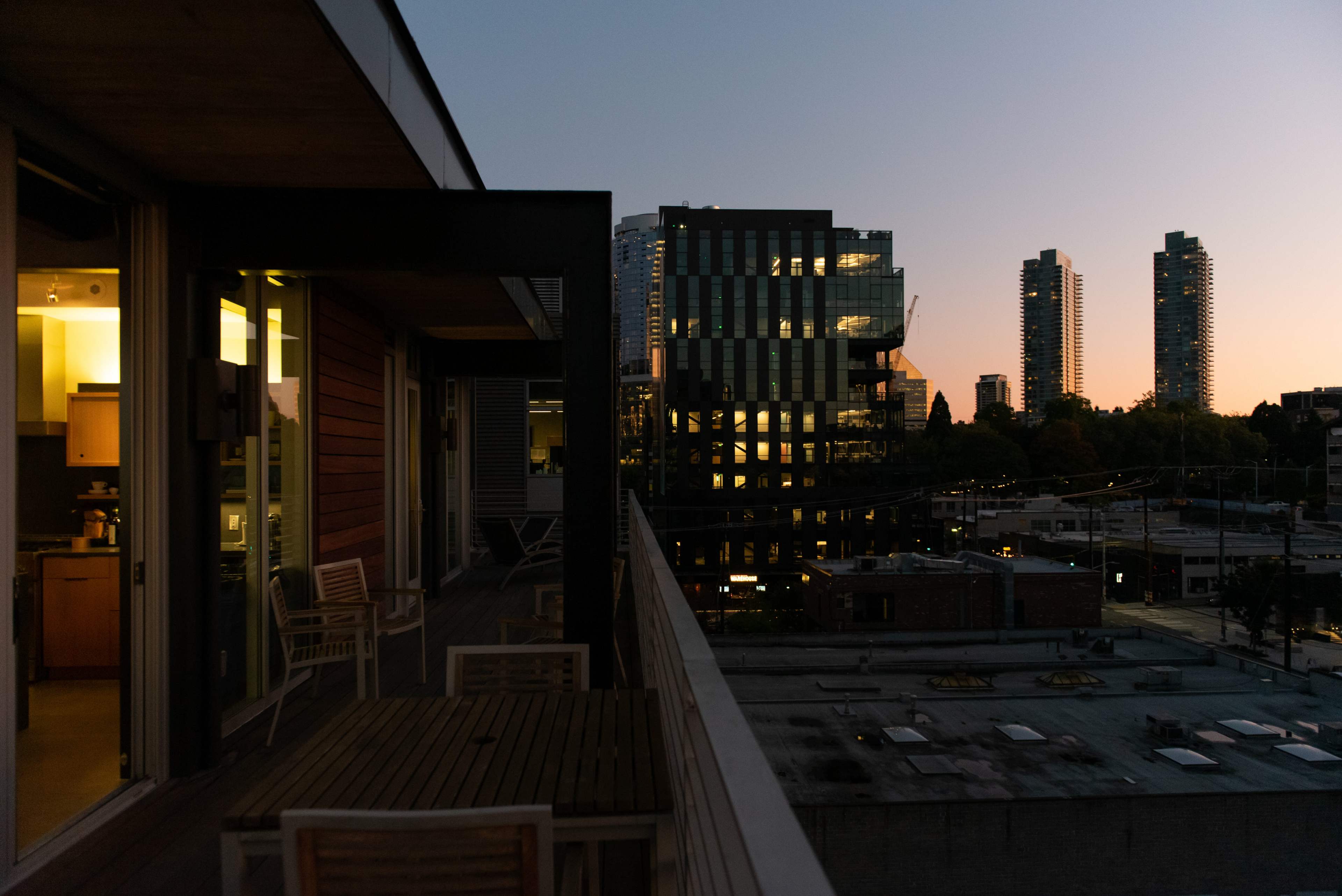The image shows a balcony with wooden furniture overlooking a city skyline at dusk, with illuminated buildings and a colorful sunset in the background.