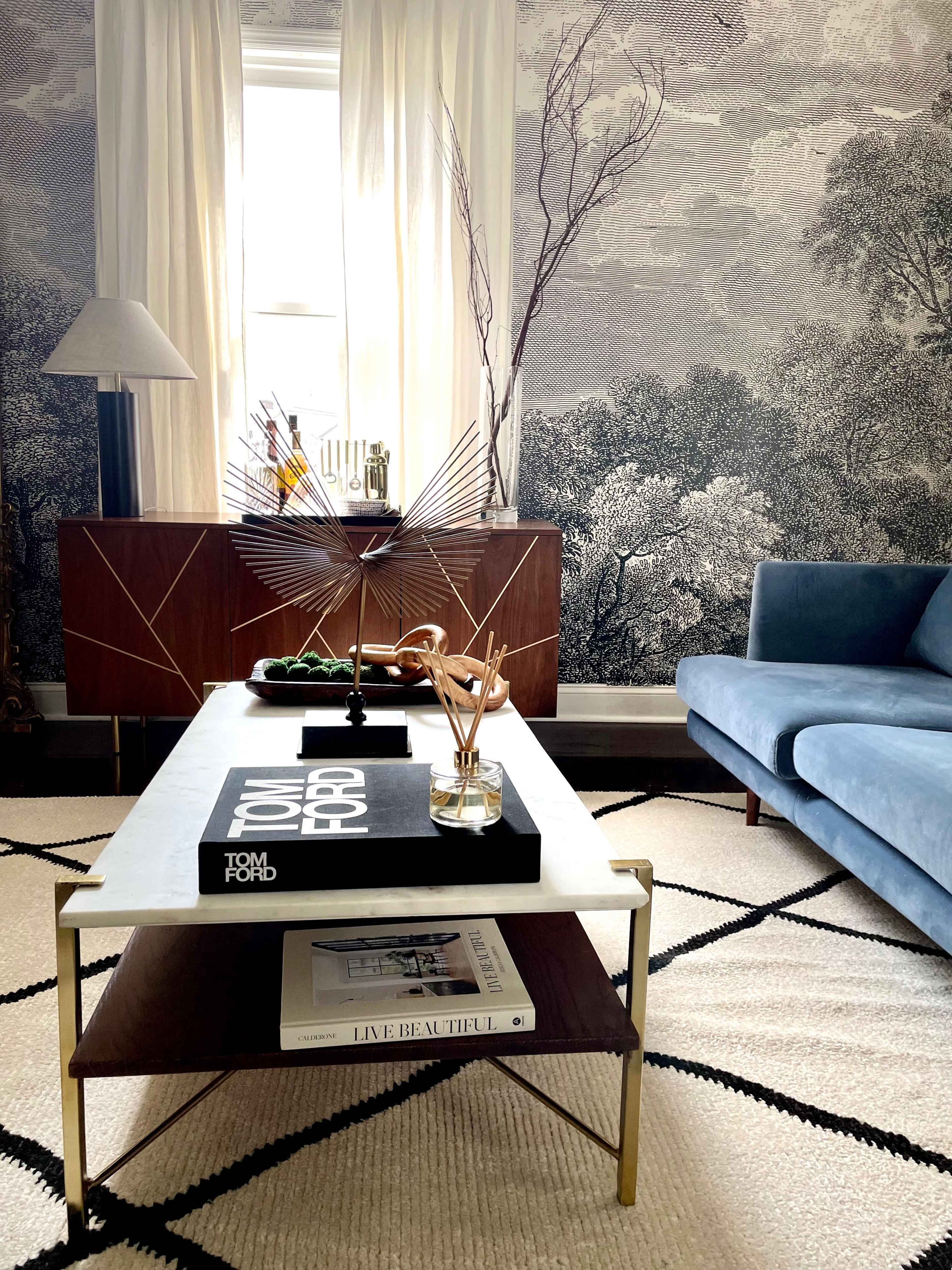 A stylish living room featuring a blue sofa, a coffee table with decorative items, and wallpaper depicting a landscape scene.